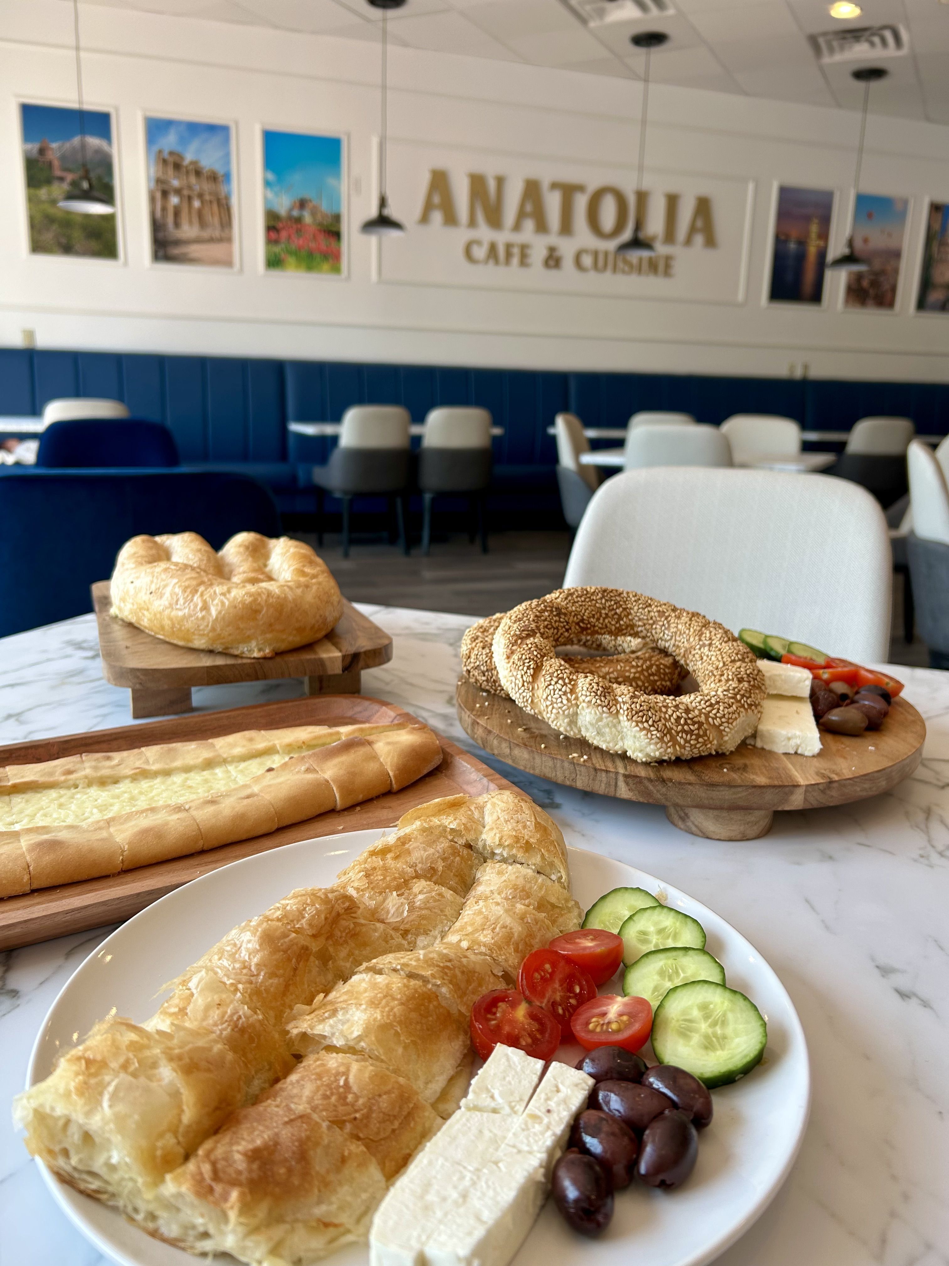 Table with Turkish breakfast including flaky pastry, sesame bagels, cucumbers, cherry tomatoes, olives, and feta cheese in Anatolia Cafe & Cuisine with blue seating and framed wall photos.