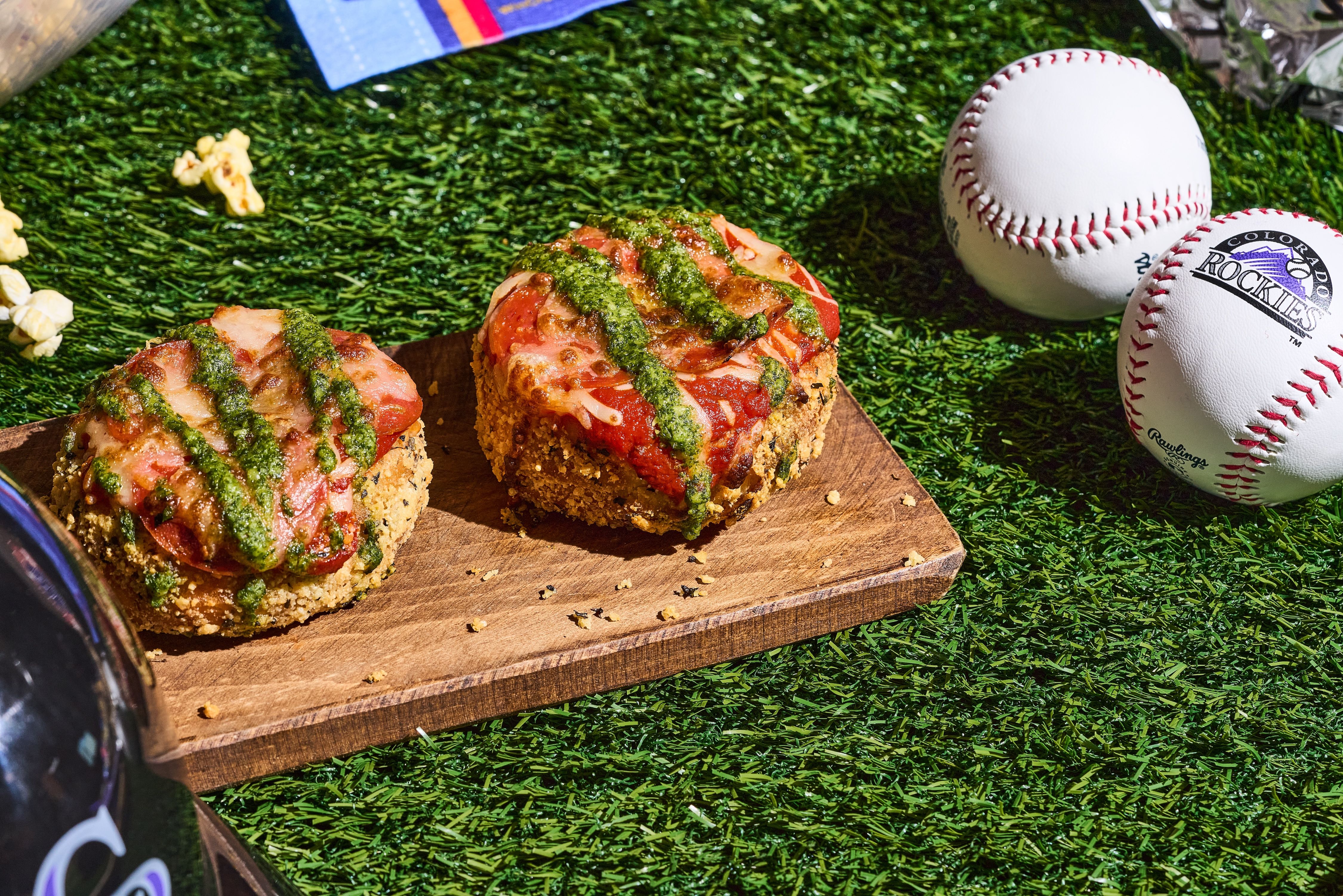 Two round, crusty sliders topped with tomato, melted cheese, and green pesto sit on a wooden board atop artificial grass, with baseballs and scattered popcorn in the background.