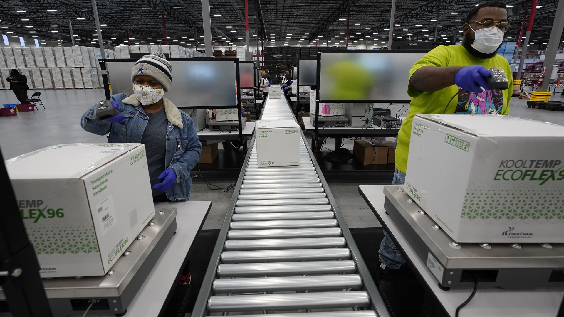 Two masked employees stand on opposite sides of an empty assembly line