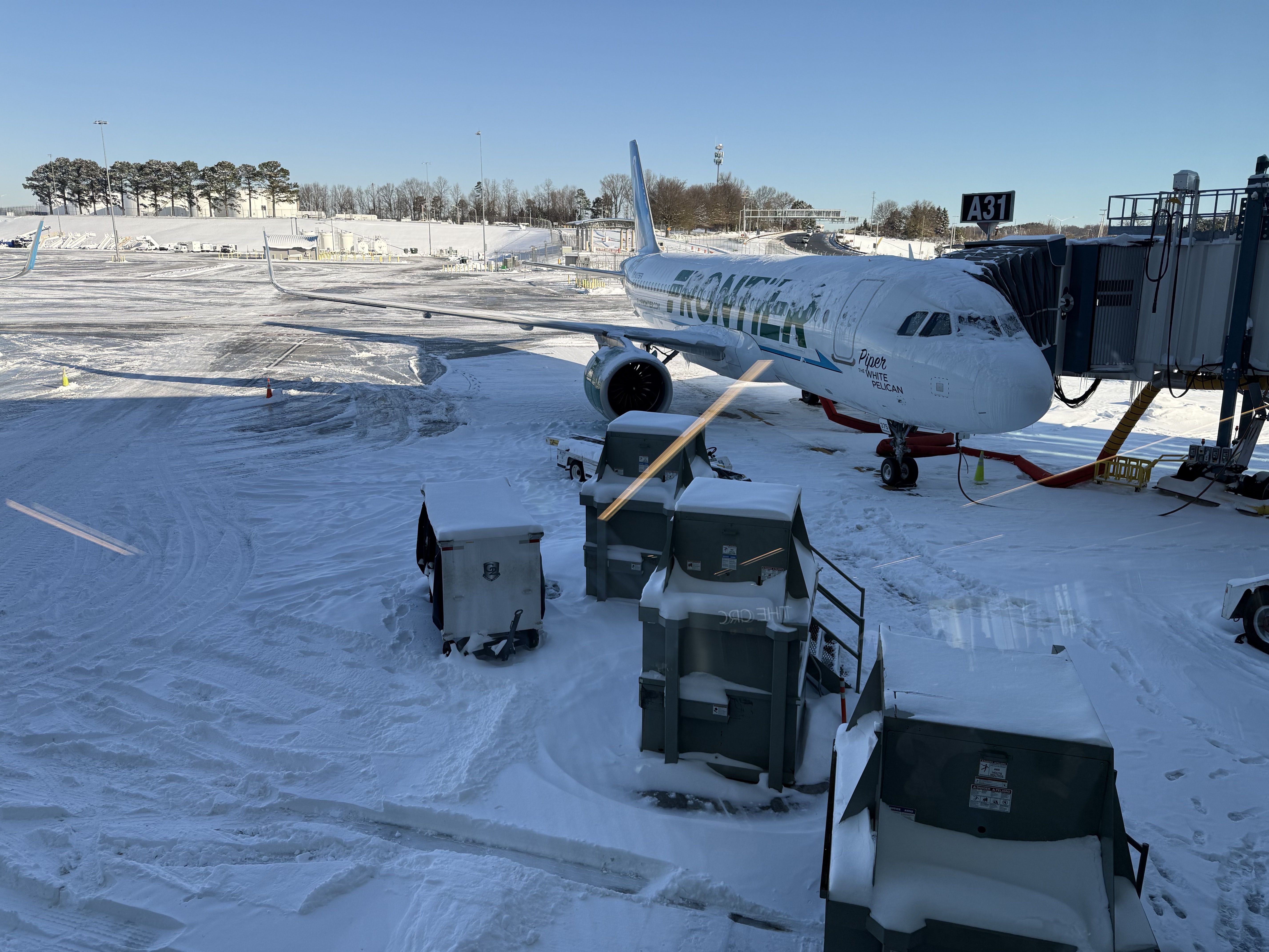 Frontier Airlines plane named Piper the White Pelican covered in snow at gate A31 with snowy airport tarmac and equipment under a clear blue sky.