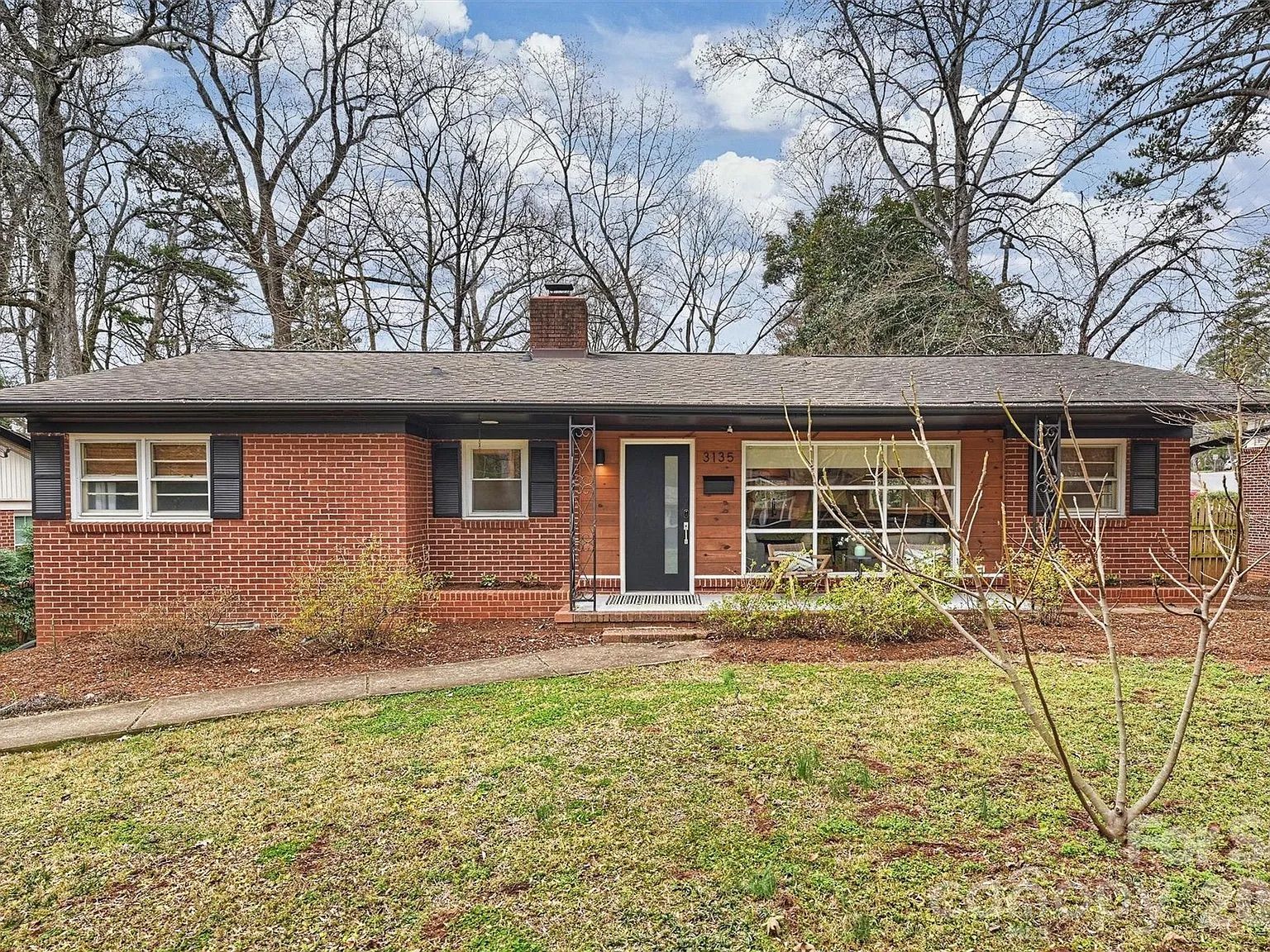 Single-story red brick house with black shutters, a black front door, large front window, leafless trees, and a green yard under a partly cloudy blue sky.