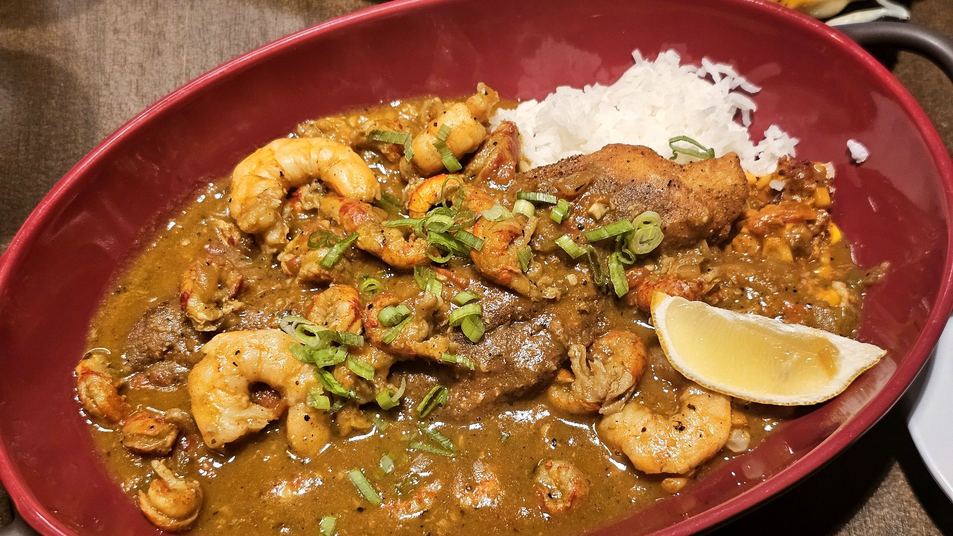 Red bowl of shrimp and beef curry served over white rice, topped with chopped green onions. Rich brown sauce coats juicy shrimp and beef pieces, with a lime wedge on the side.
