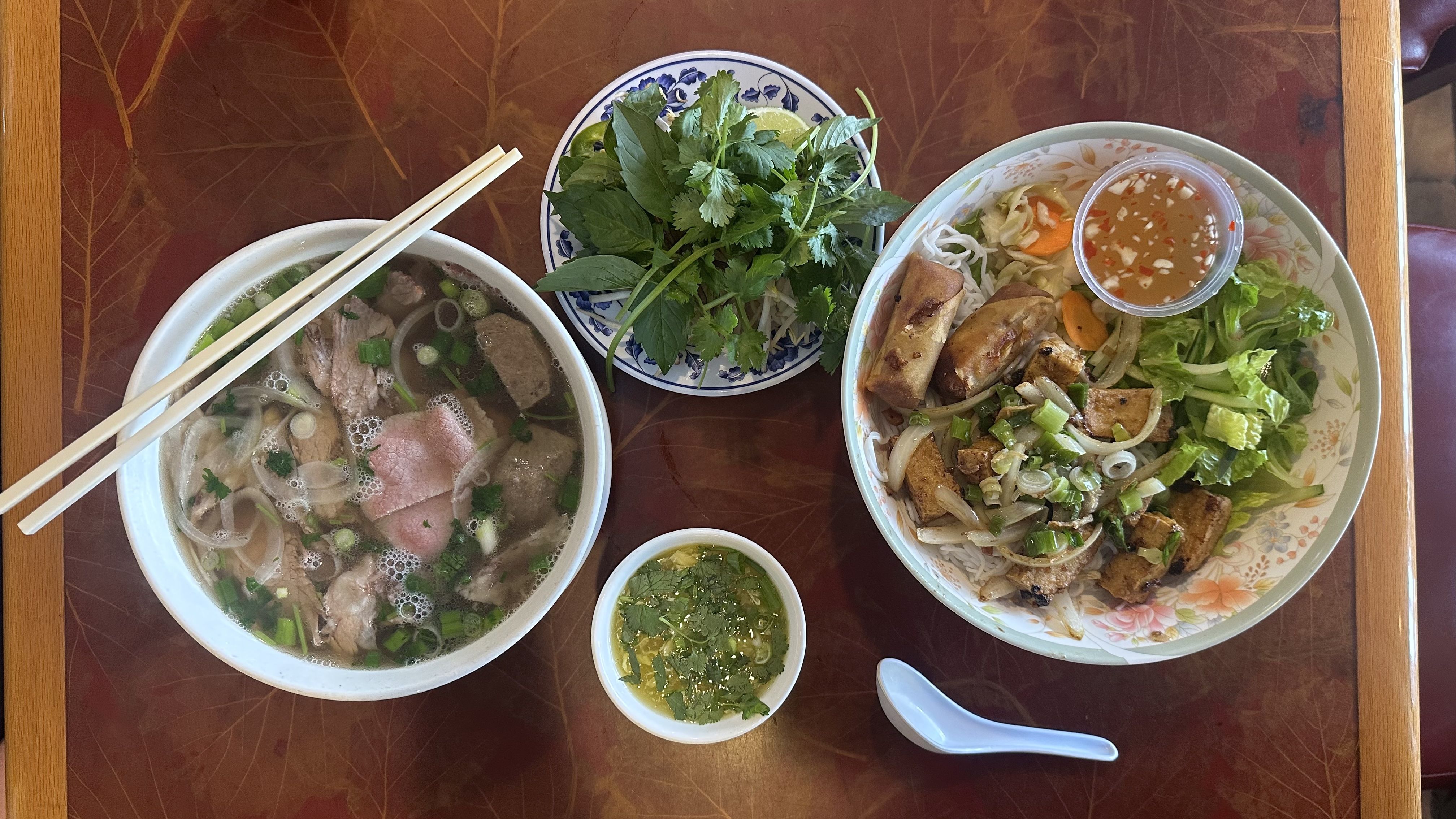 Vietnamese meal with a bowl of pho containing meat and green onions, a plate of fresh herbs and lime, a bowl with tofu, veggies, and dipping sauce, all on a wooden table.