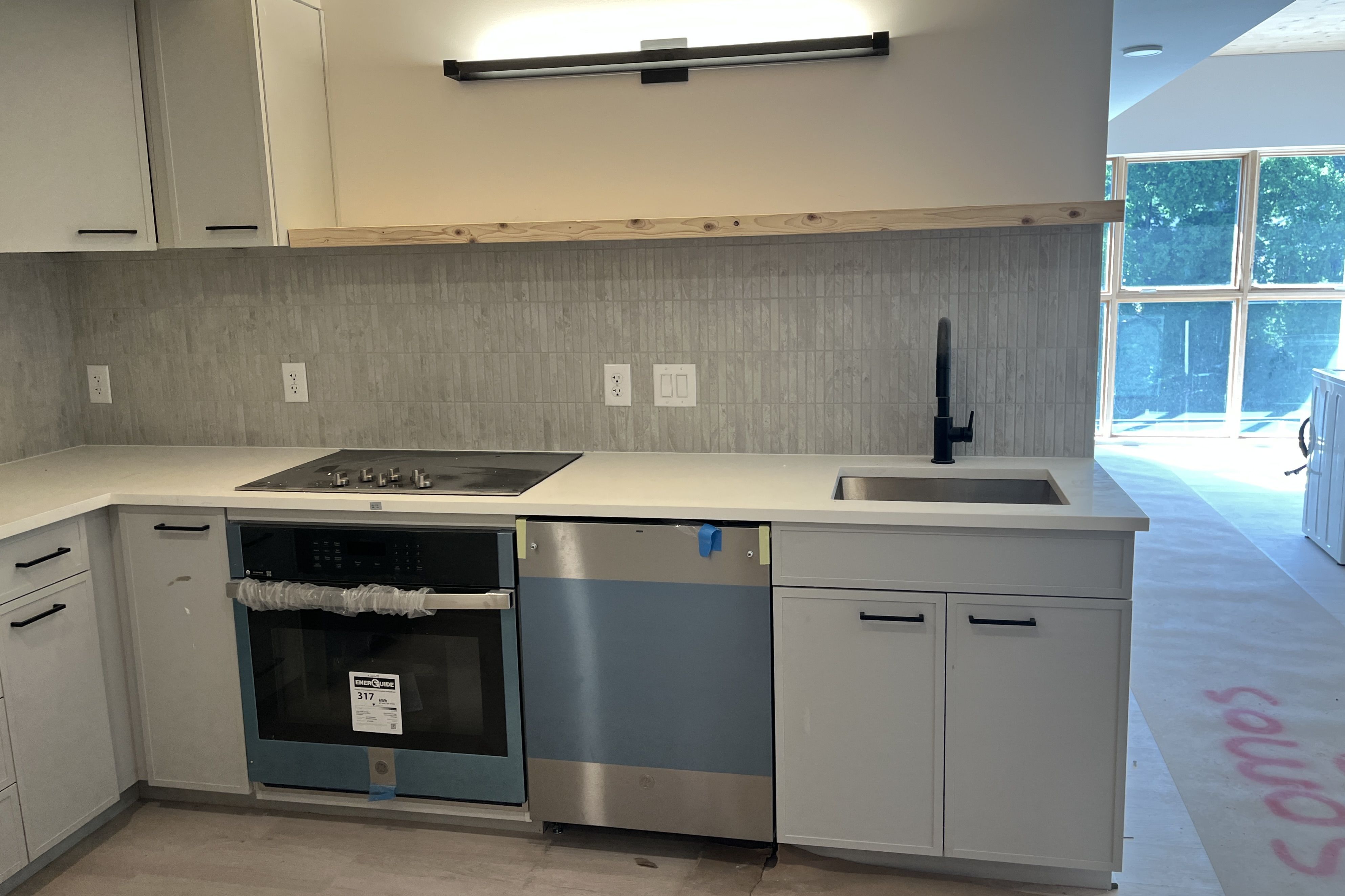Modern kitchen with gray cabinets, vertical tile backsplash, stainless oven and dishwasher, black faucet over a white sink, light wood trim along the wall, and windows with greenery outside.