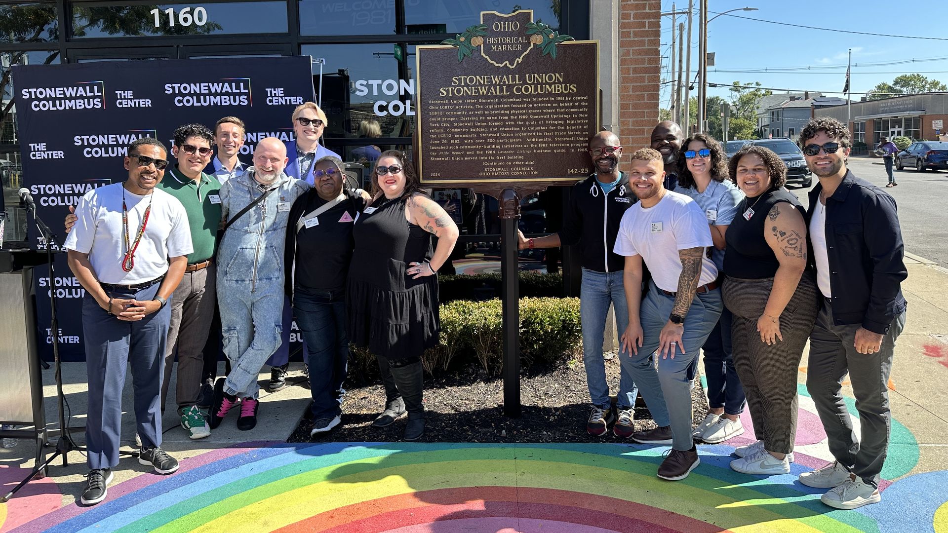 Group posing outside a brick building with a rainbow crosswalk and Stonewall banners, beside a large Ohio Historical Marker about Stonewall Union/Stonewall Columbus.