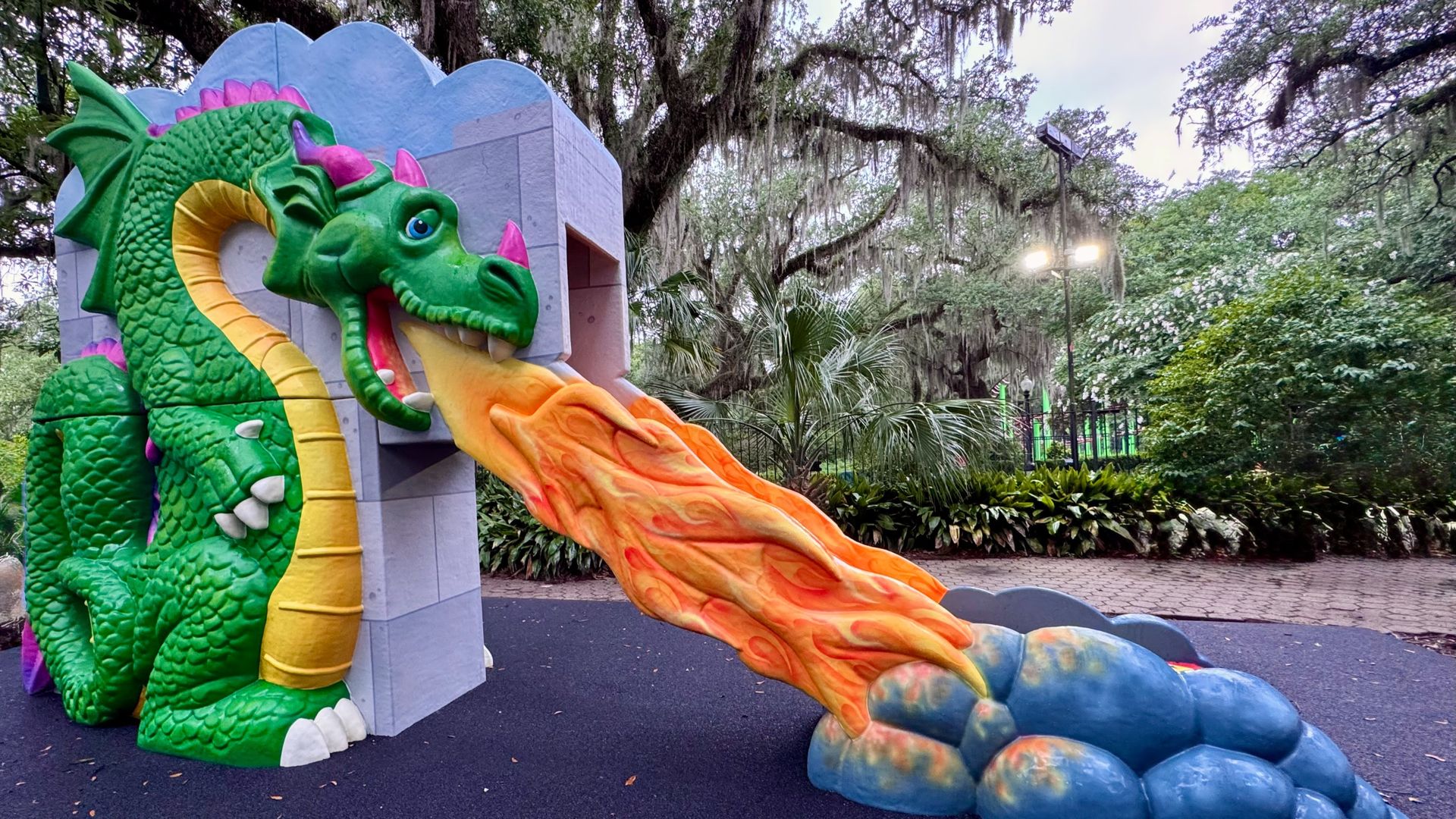 Bright green dragon sculpture at a park slide, yellow belly, pink spikes, blue eyes, spouting an orange flame slide from a gray stone entrance, with leafy trees in the background.