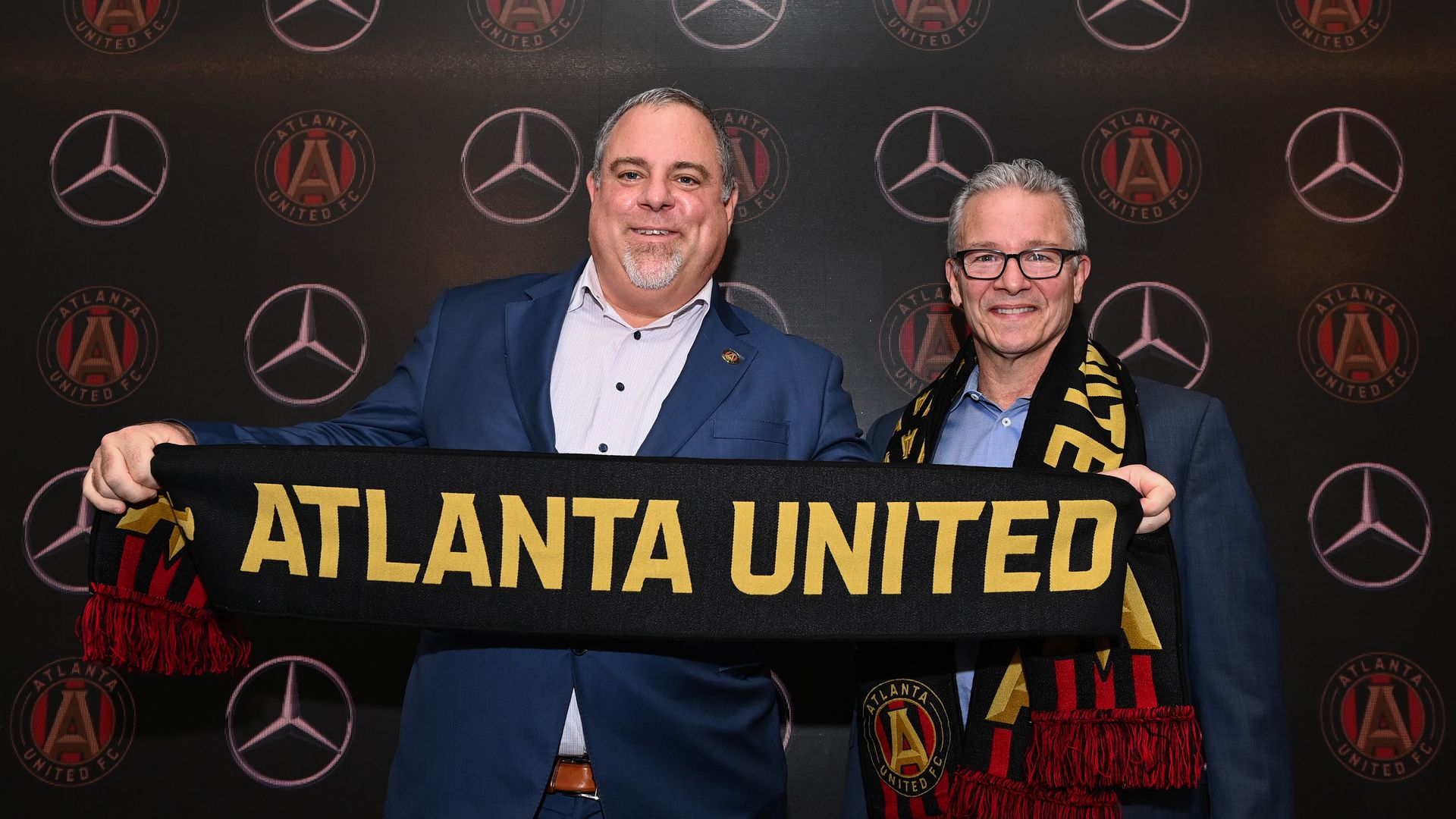 A man in a suit poses with another man while he holds a scarf that says "Atlanta United" 
