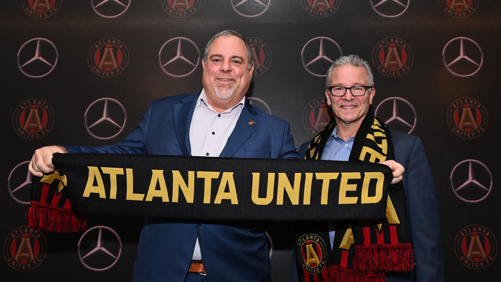 A man in a suit poses with another man while he holds a scarf that says "Atlanta United"