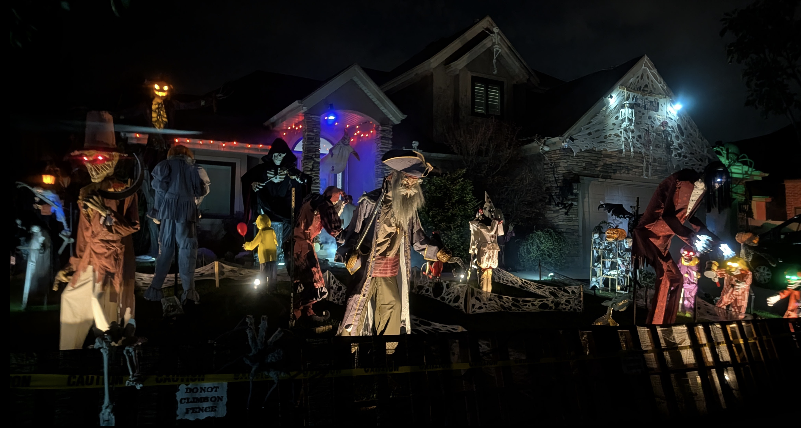 Nighttime Halloween yard scene with life-size skeletons and eerie figures, orange and blue lights, a carved pumpkin, spider-web garlands on the house, and caution tape in the foreground.