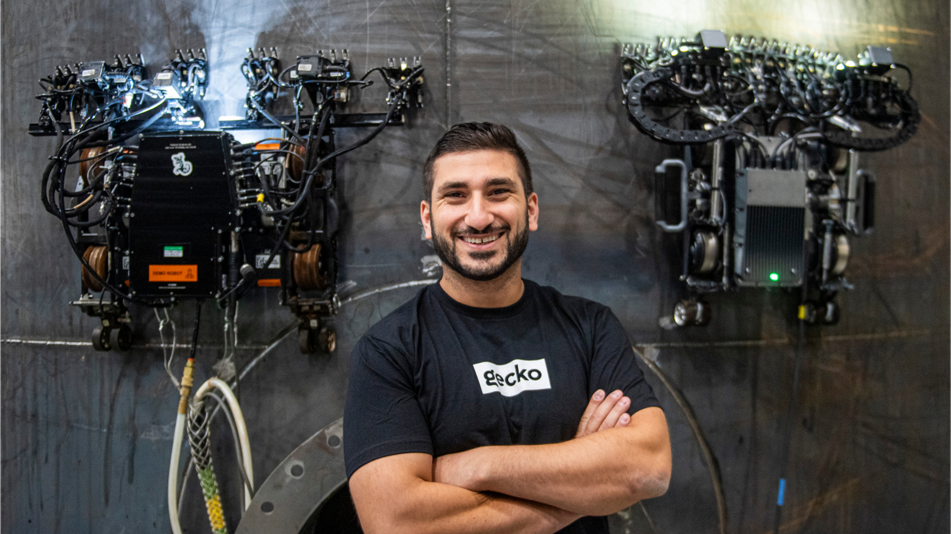 A man stands with his arms folded across his chest. He is flanked by two wall-climbing robots.