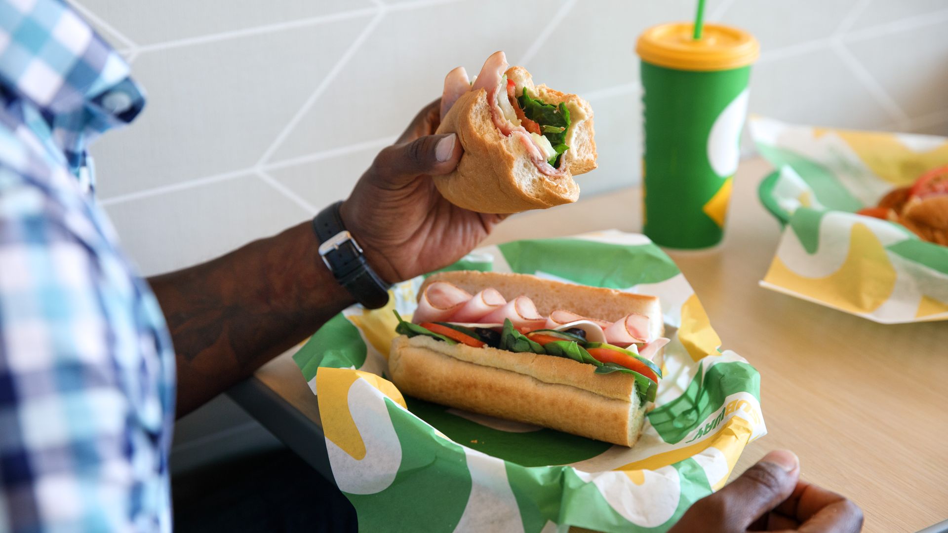 Hands holding half of a sandwich and the other half on a wrapper. Cup in background