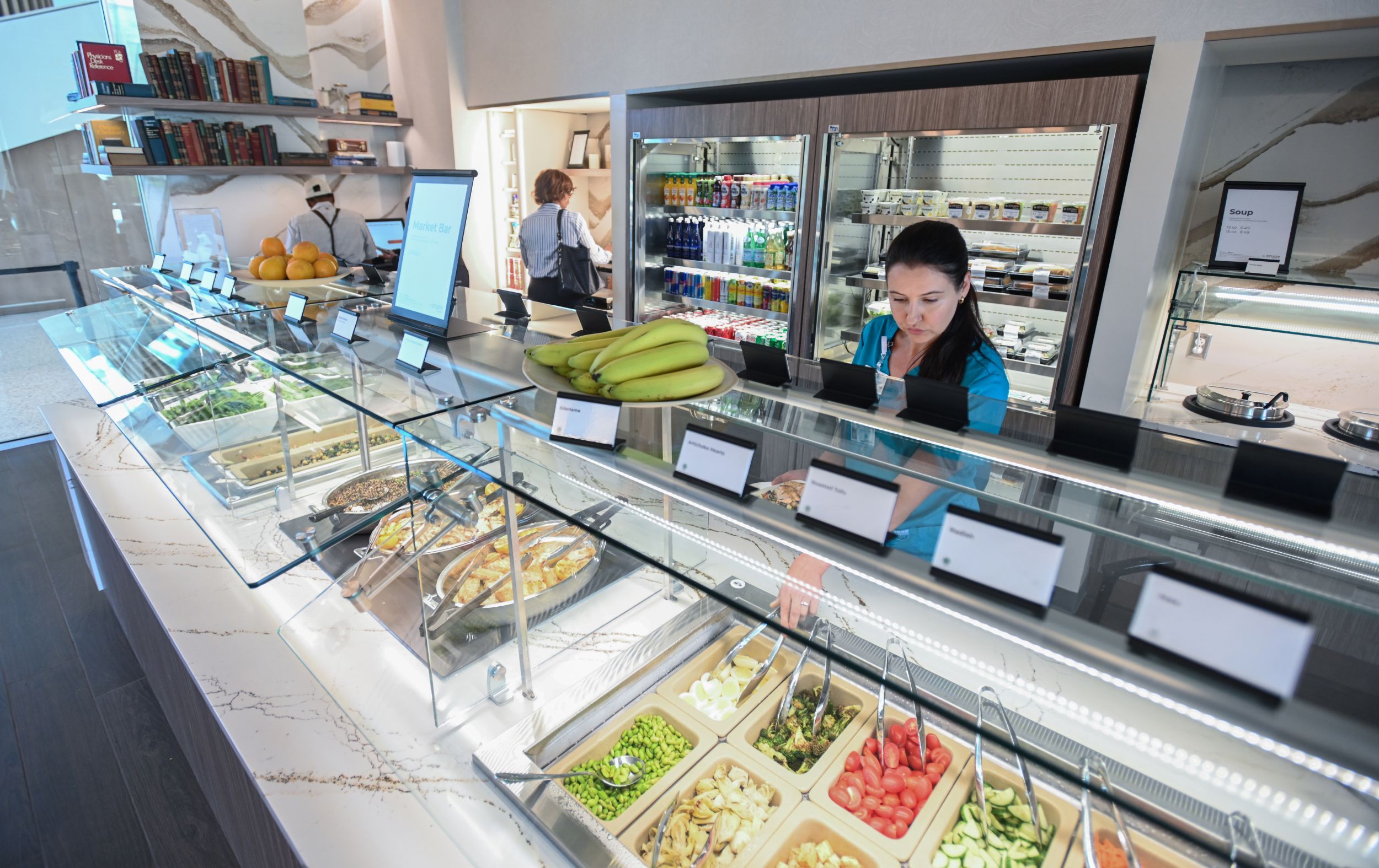 Modern cafe with glass display cases of salads and prepared foods, bananas on the counter, a woman in teal behind the counter, and refrigerated shelves in back; books line the left wall.