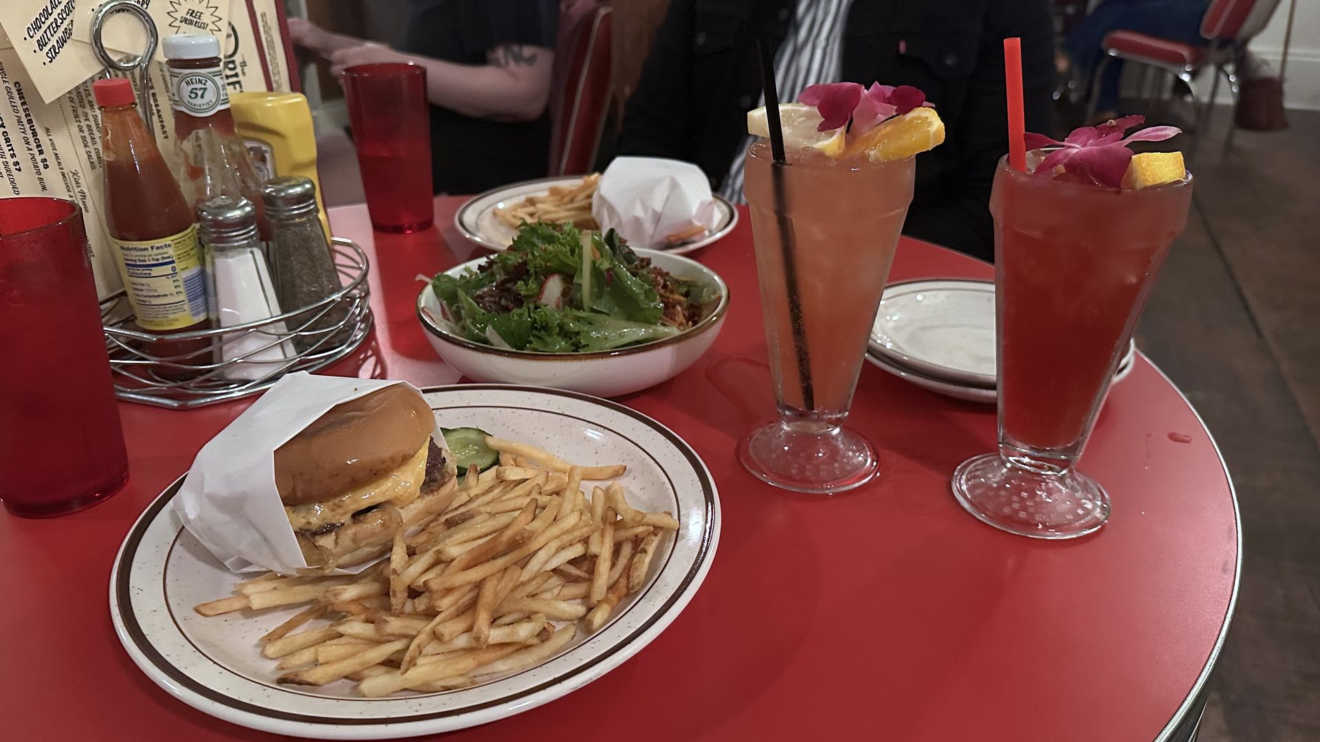 Red table with plates of cheeseburgers, fries, salad, and two colorful drinks garnished with orange slices and purple flowers, condiments and red cups in the background.