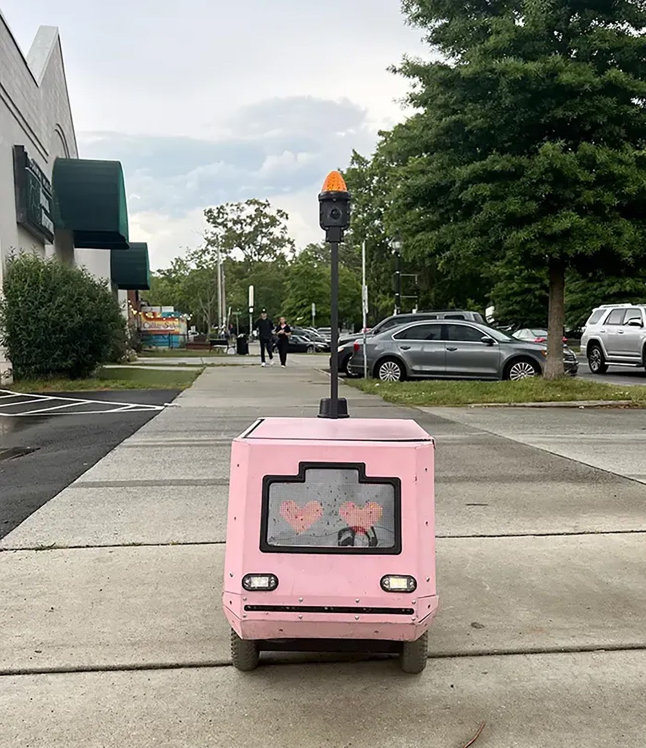 Small pink delivery robot on a sidewalk with a screen displaying two red hearts, an orange light on a pole, parked cars, and two people walking in the background.
