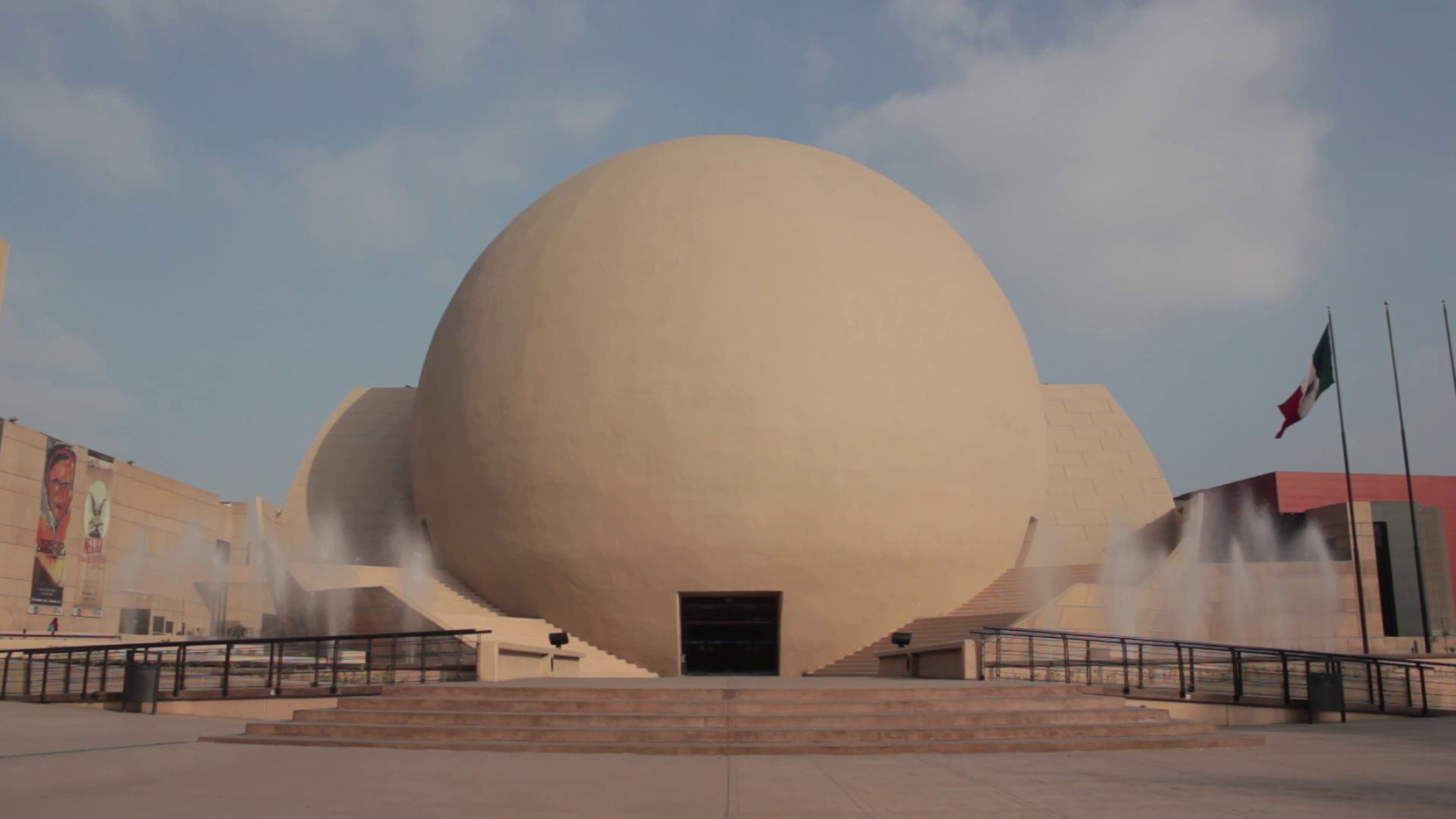 Wide view of a large beige sphere-shaped building with steps and fountains at the entrance; a Mexican flag on flagpoles stands to the right under a blue sky with clouds.