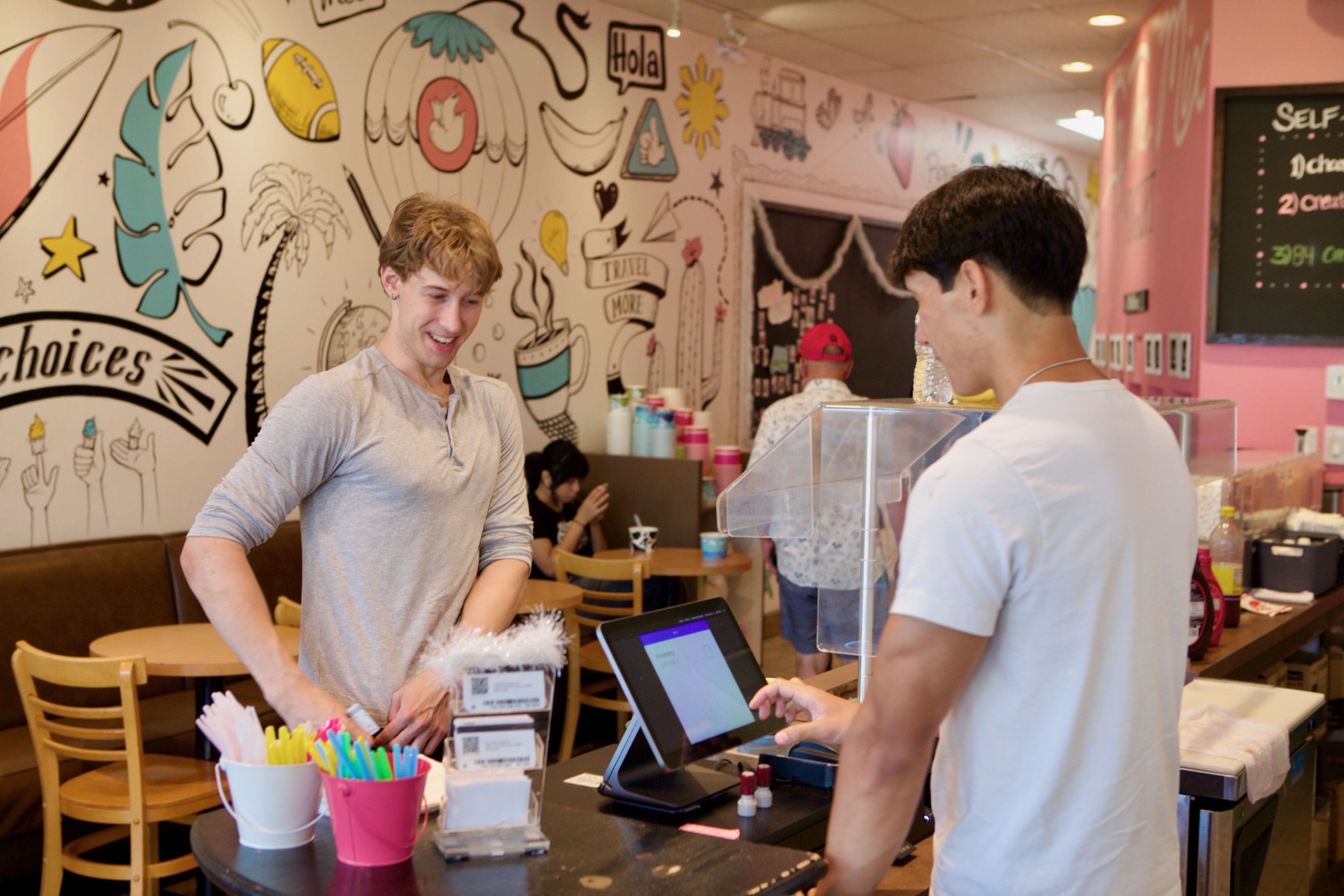 Two young men stand at a cafe counter with a colorful doodle mural; one smiles in a gray shirt, the other faces the register. Cups with straws sit in front.