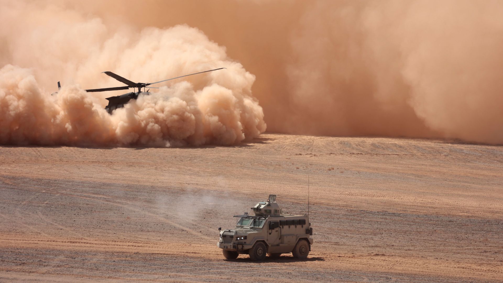 Desert scene with a military helicopter stirring up a large orange dust cloud; an armored vehicle sits in the foreground as sand storms rise in the distance.