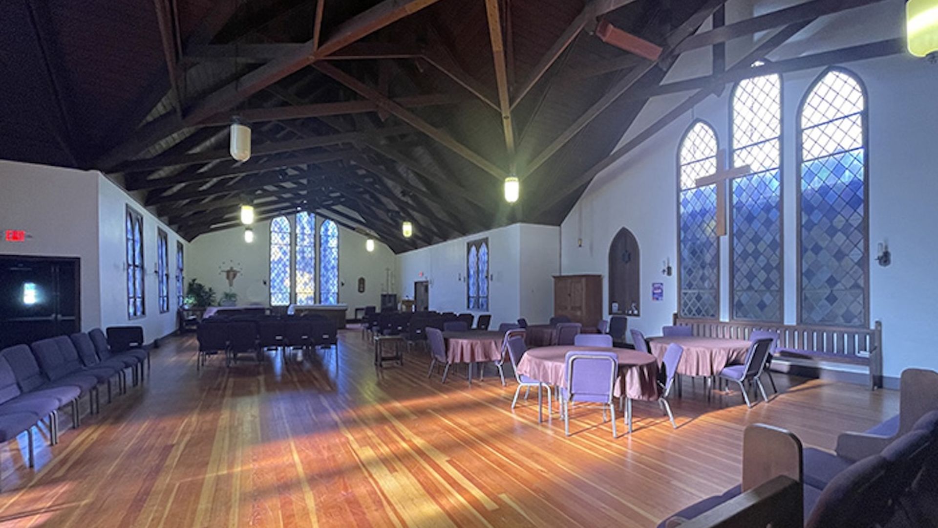 The inside of a church sanctuary shows hardwood floors, tall ceilings and tables and chairs.