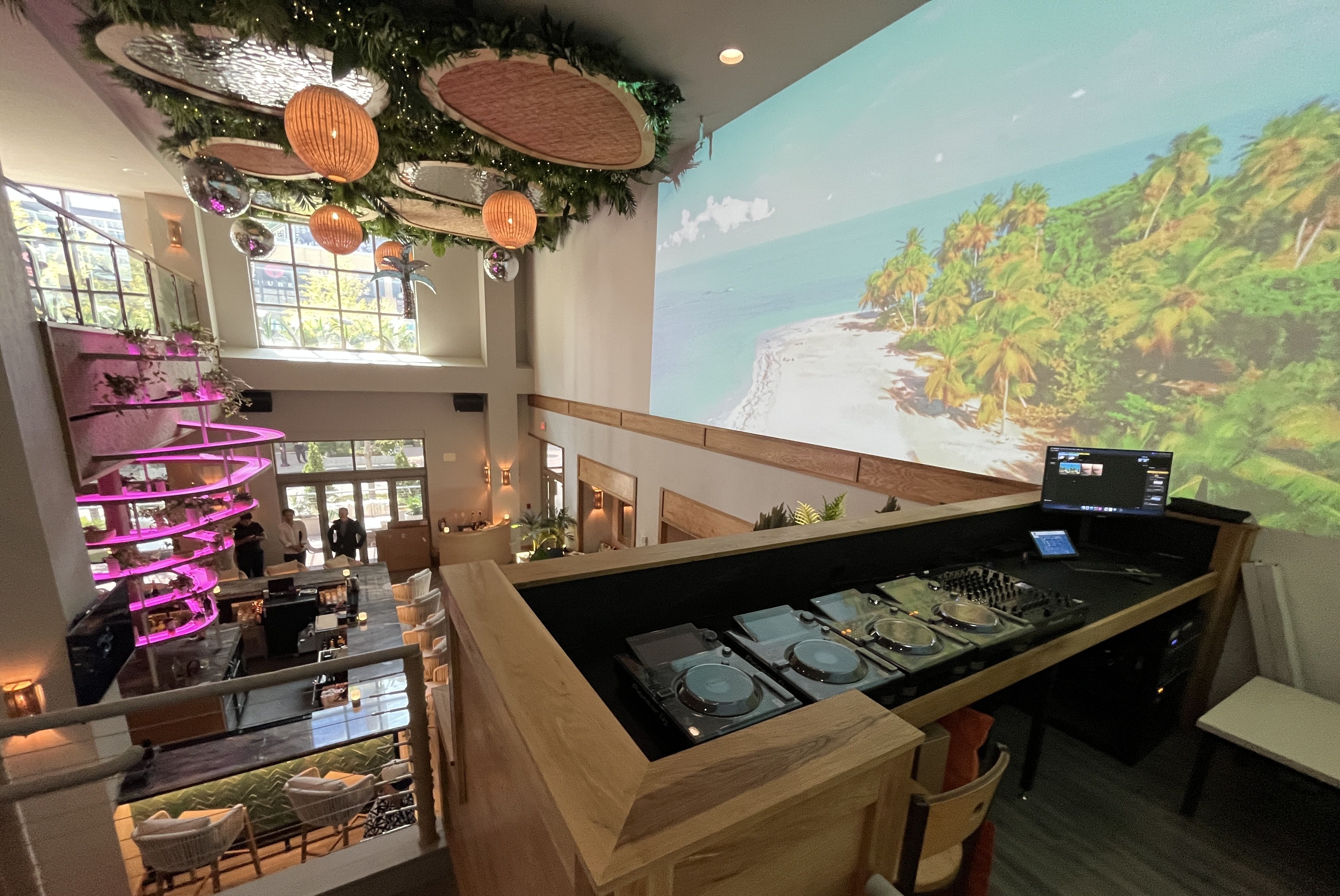 Interior of a modern lounge with a DJ booth, mixing equipment, a large beach projection on the wall, hanging wicker lights, and a pink-lit bar with patrons near windows.