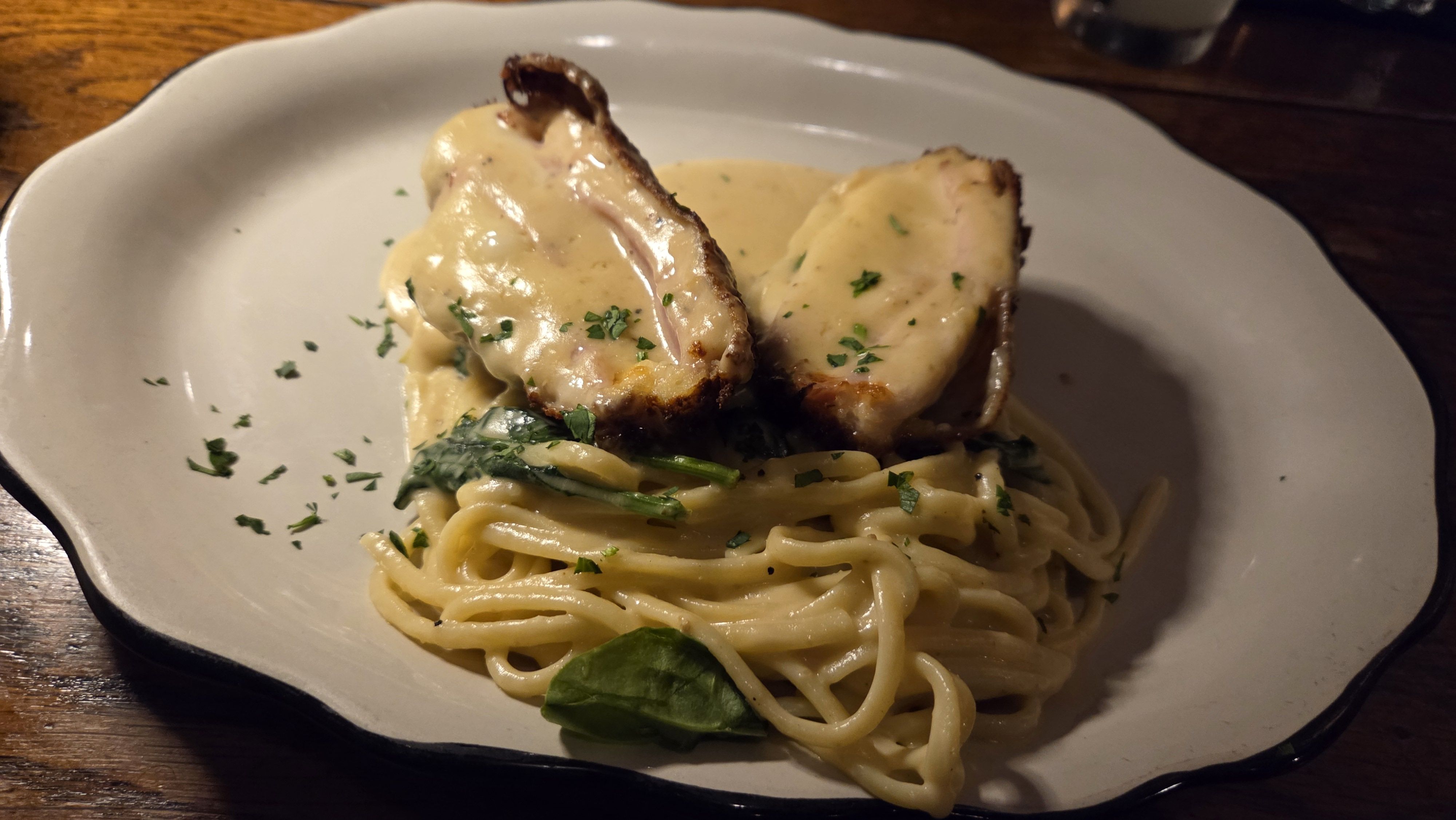 White plate with creamy pasta topped with sautéed spinach and two pieces of chicken breast covered in a light yellow sauce, sprinkled with green herbs, on a wooden table.
