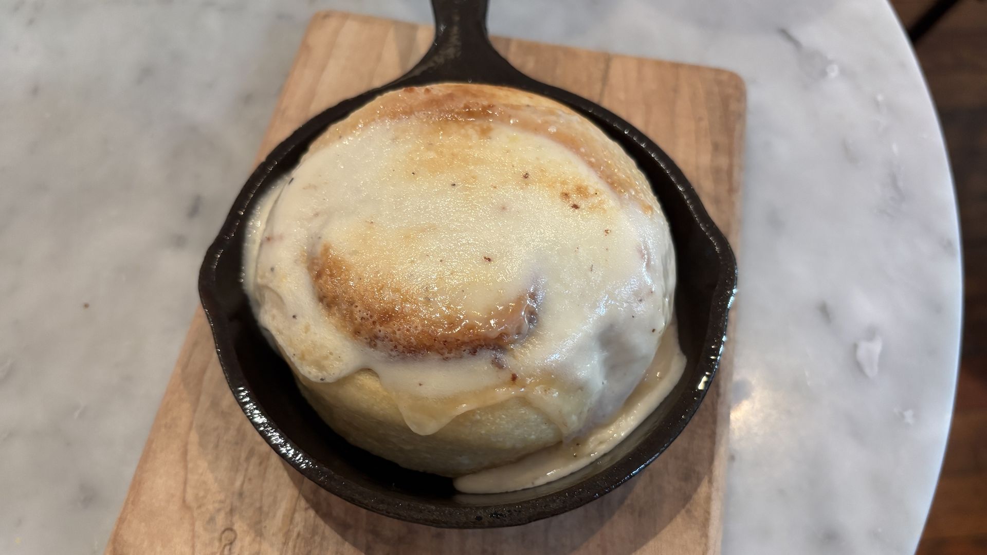 A cinnamon bun with melted white icing sits in a small black cast-iron skillet on a wooden board, atop a light marble counter.