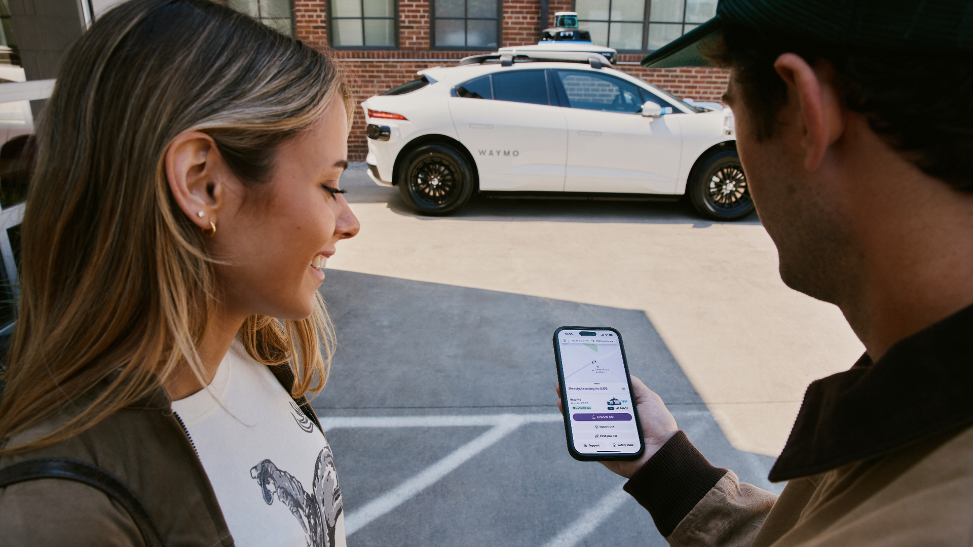 A woman and a man stand by a white Waymo self-driving car with roof sensors, parked in front of a brick building; They are looking at the Lyft app on the man's phone.