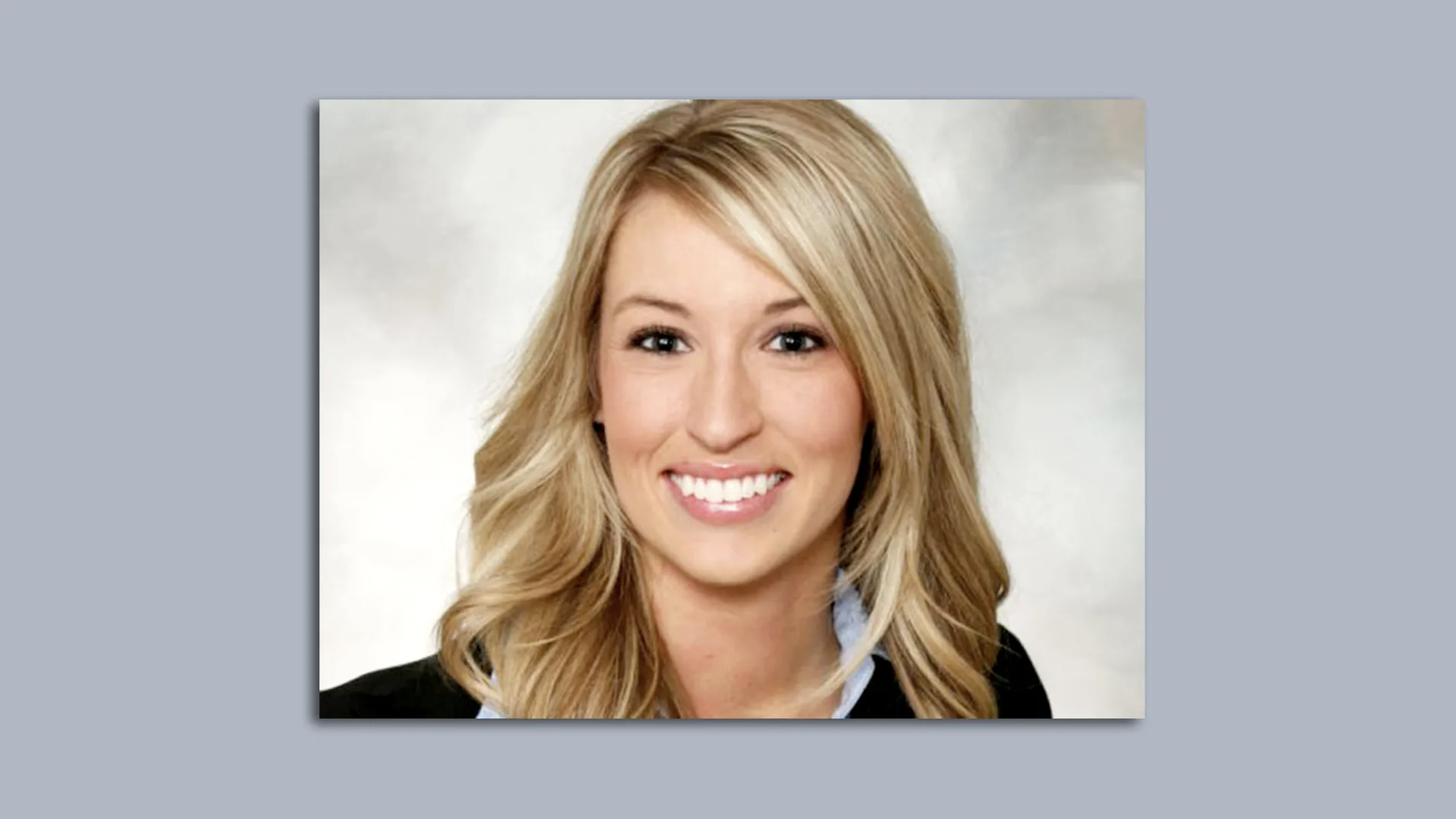 Portrait of a smiling blonde woman with wavy hair, wearing a dark blazer over a light blue shirt, against a light gray studio backdrop.