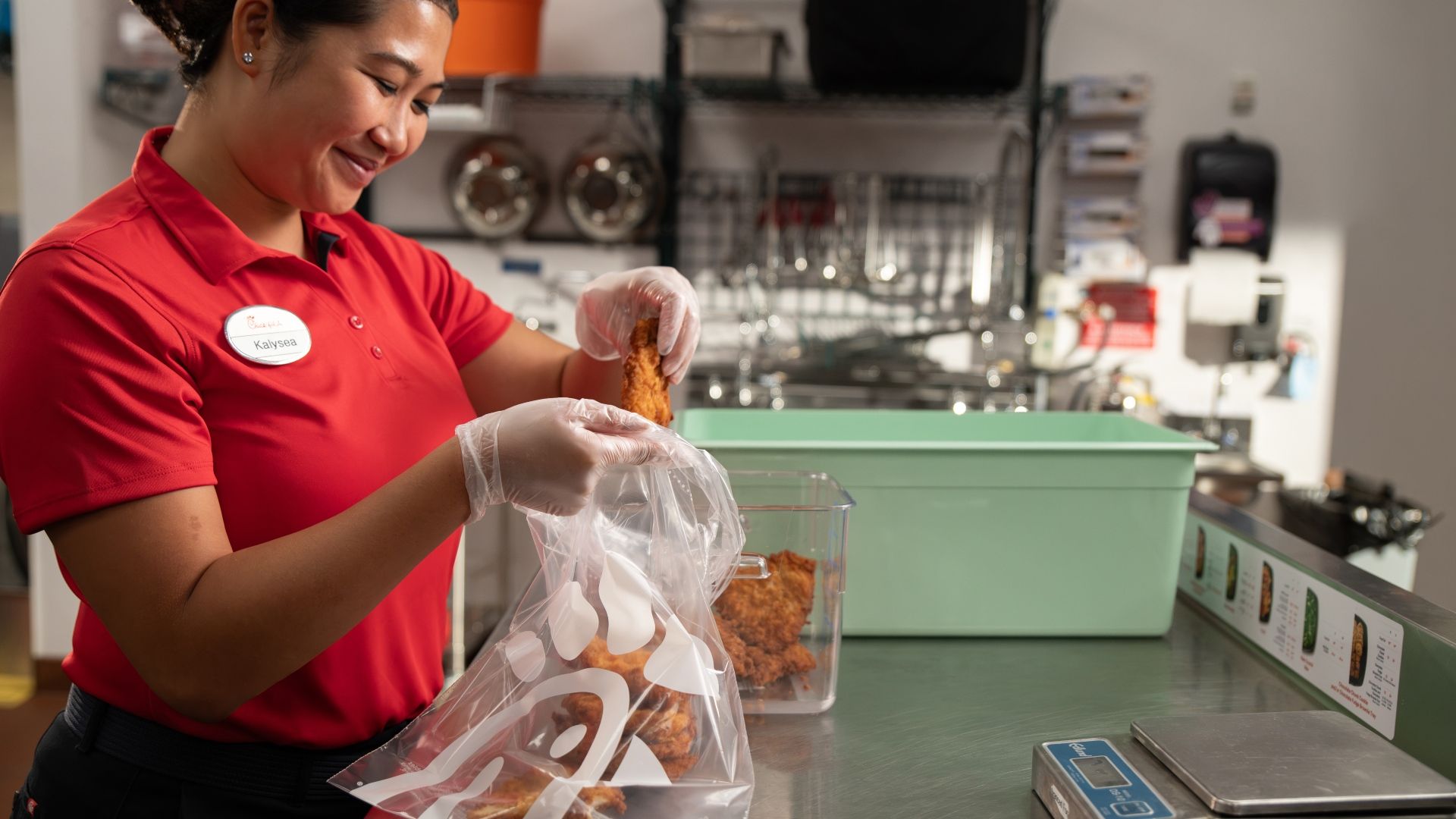 A smiling food worker in a red polo shirt bags fried chicken into a clear plastic bag, in a kitchen with metal shelves, a green bin, and a scale on the counter.