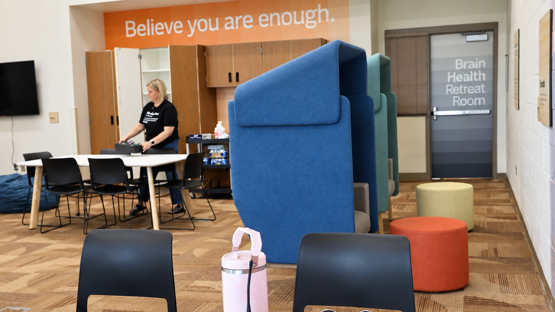 Bright room with an orange wall that says Believe you are enough. A woman in a black shirt sorts items at a table; blue modular seating and colorful ottomans line the area, near a Brain Health Retreat Room door.