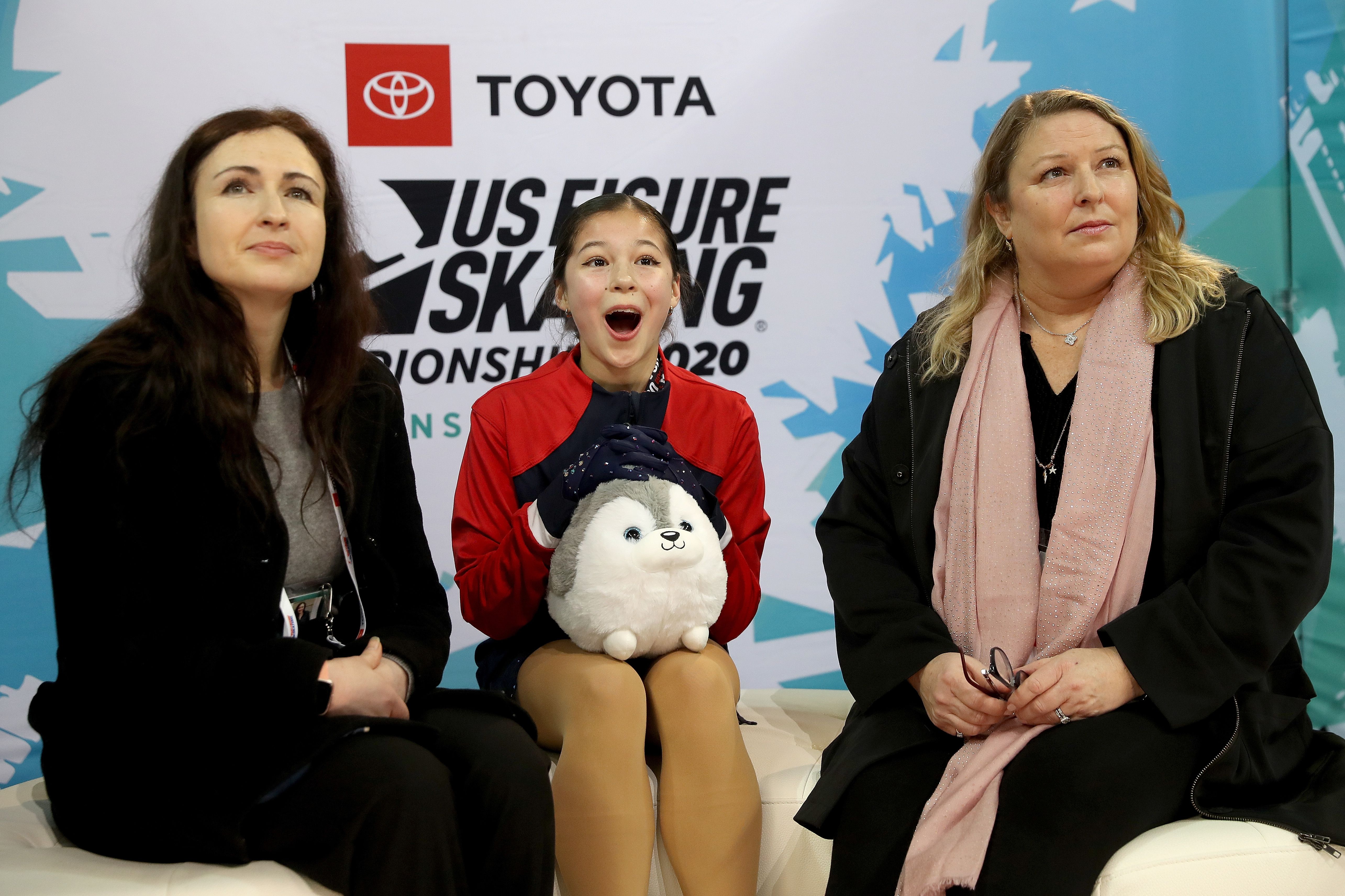 Three women seated against a US Figure Skating Championship 2020 backdrop. The young woman in the center wears a red jacket and holds a white-and-gray plush toy, looking excited. The other women wear black.