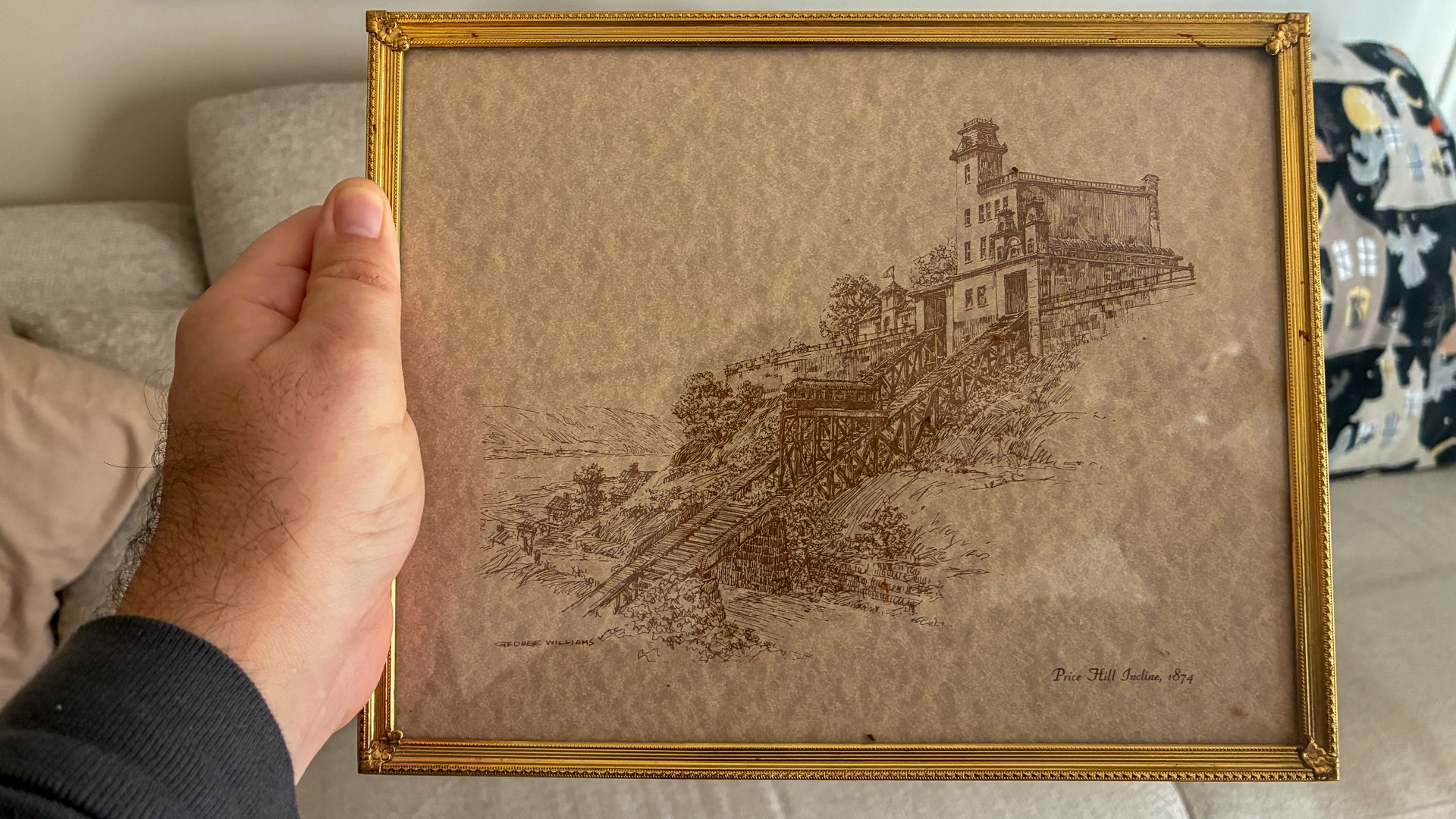 Hand holds a gold-framed brown sheet featuring a detailed ink engraving of a hillside incline with buildings and a bridge; reads 'George Williams' and 'Price Hill Incline, 1874'.