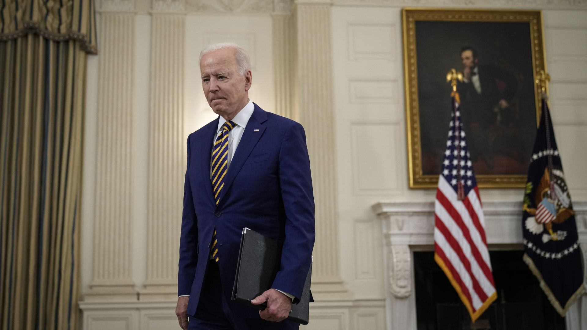 Photo of Joe Biden in a blue suit walking with his head bowed and a black binder in one arm