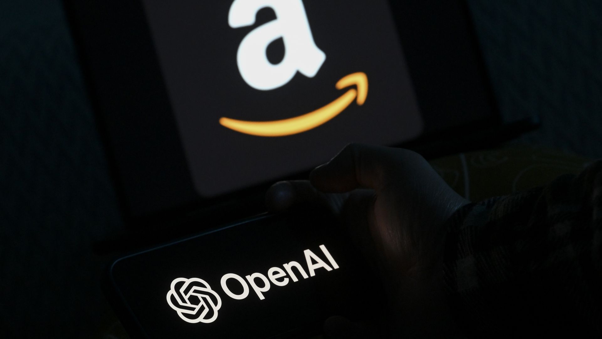 A phone displaying the OpenAI logo; in the background a tablet shows the white Amazon logo with the orange curved arrow.