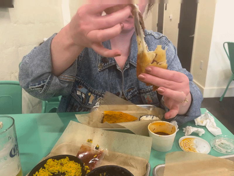 A smiling woman in a denim jacket at a teal table pulls apart an eggroll with melted cheese in the center. 
