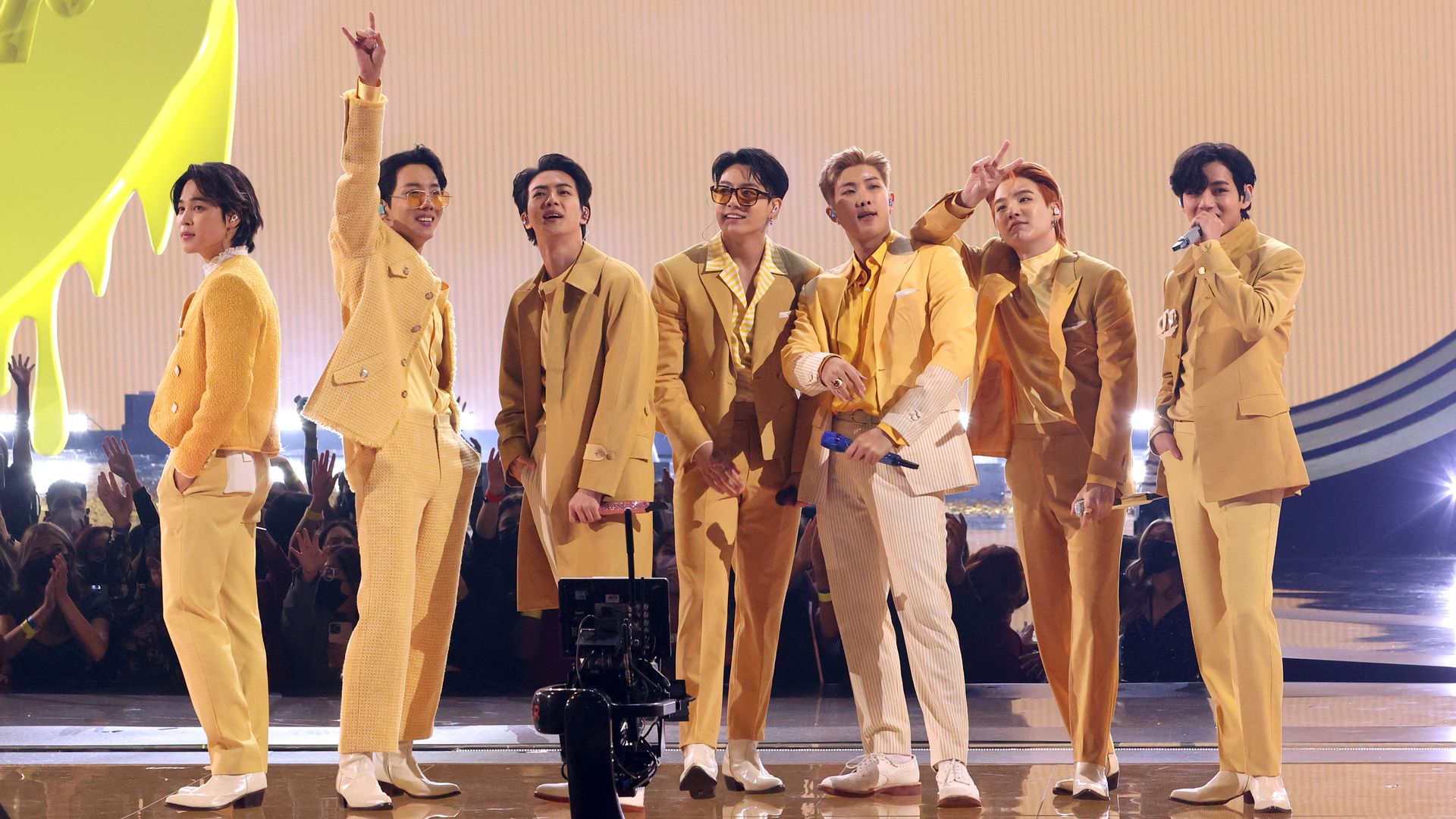 Seven men in coordinated yellow and beige outfits stand on a stage with a bright backdrop, posing and holding microphones, with an audience cheering in the background.