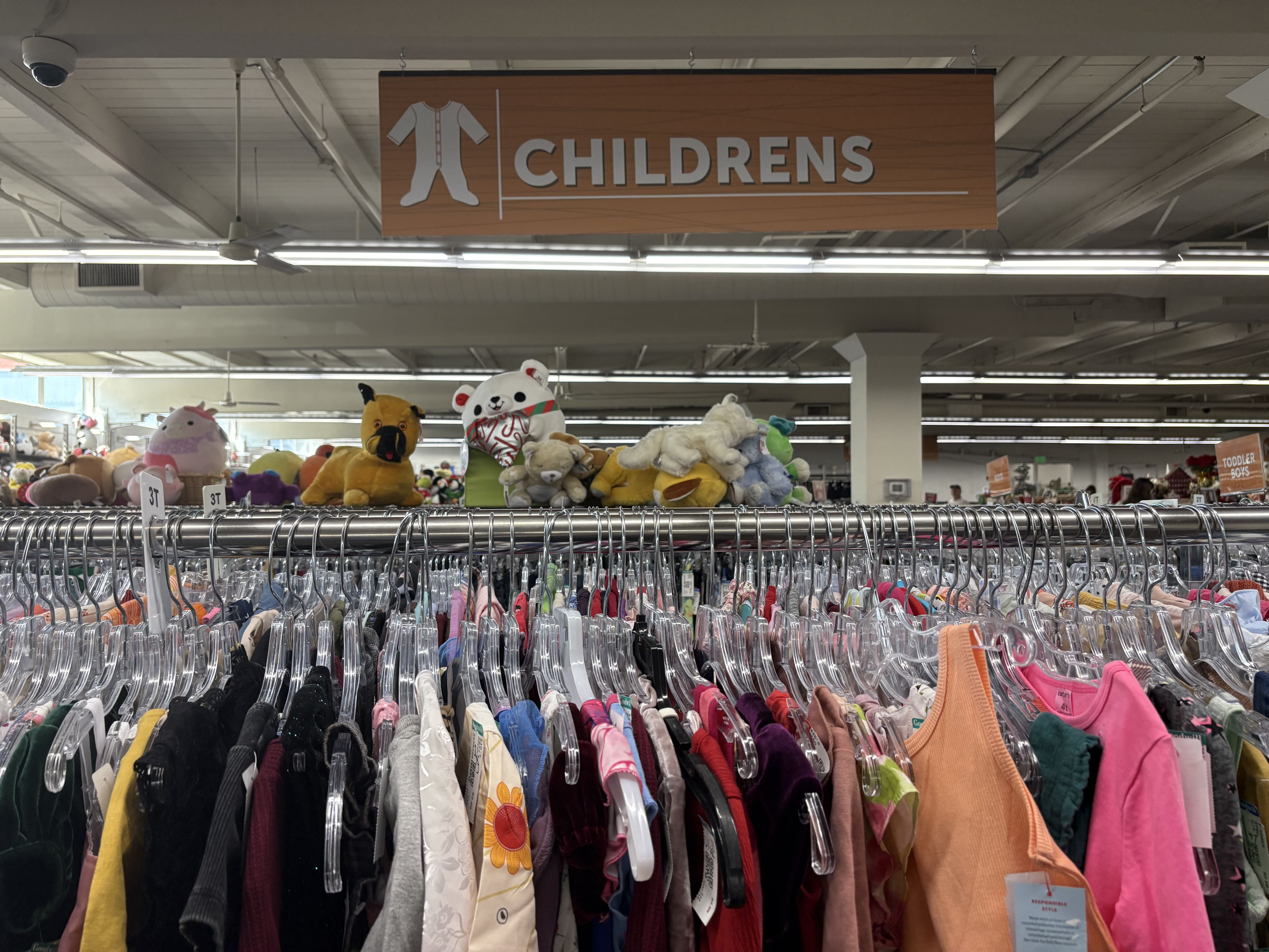 A store aisle with an orange sign reading "CHILDRENS" and a white onesie icon, hanging above racks of clothing and shelves full of stuffed animals in the background.