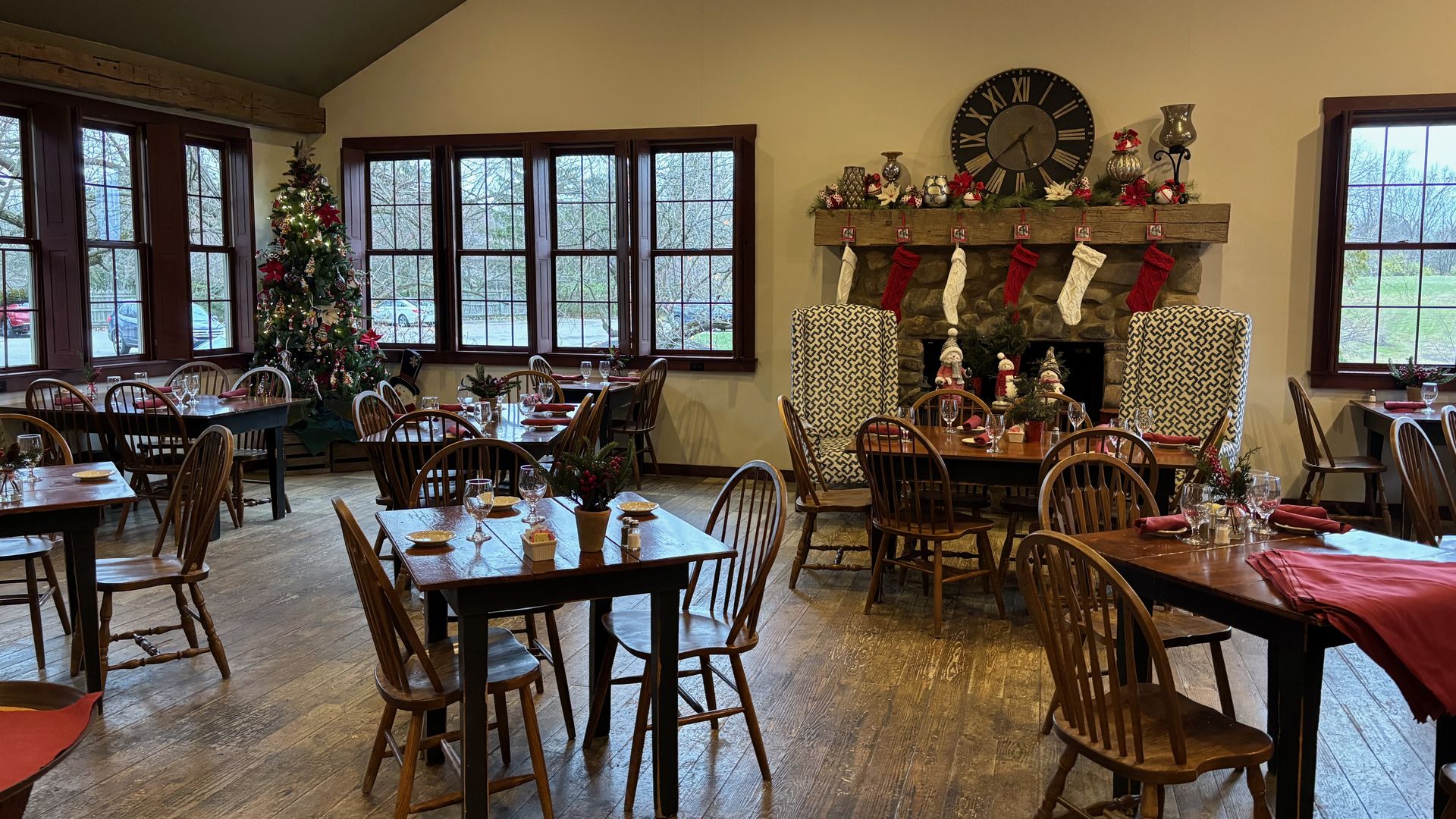 A cozy restaurant with wooden tables and chairs, a decorated Christmas tree near windows, a stone fireplace with red and white stockings, holiday decor, and a large clock above it.