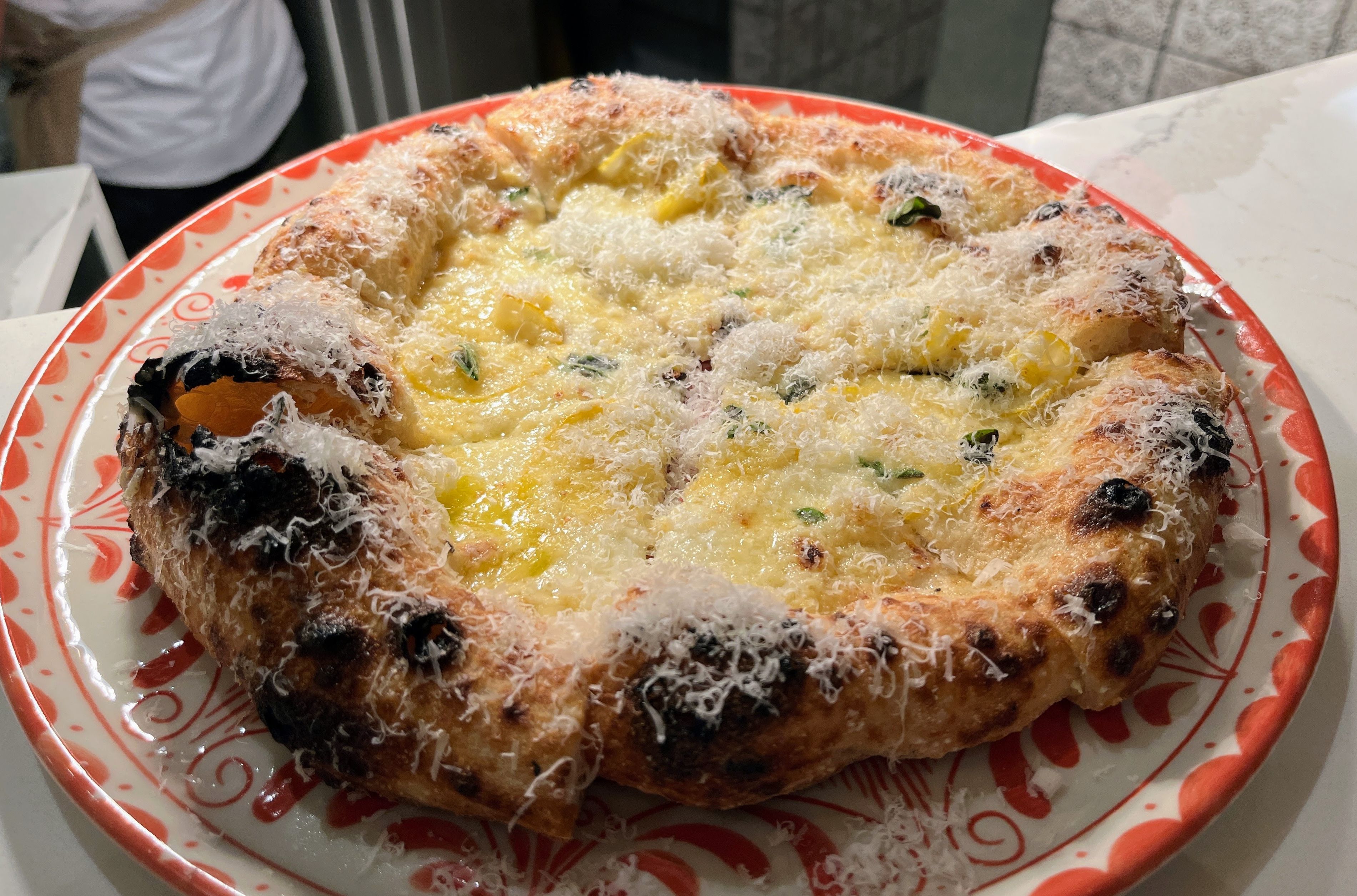 Close-up of a freshly baked pizza with a thick, charred crust, topped with melted cheese, herbs, and shredded Parmesan on a red and white patterned plate.