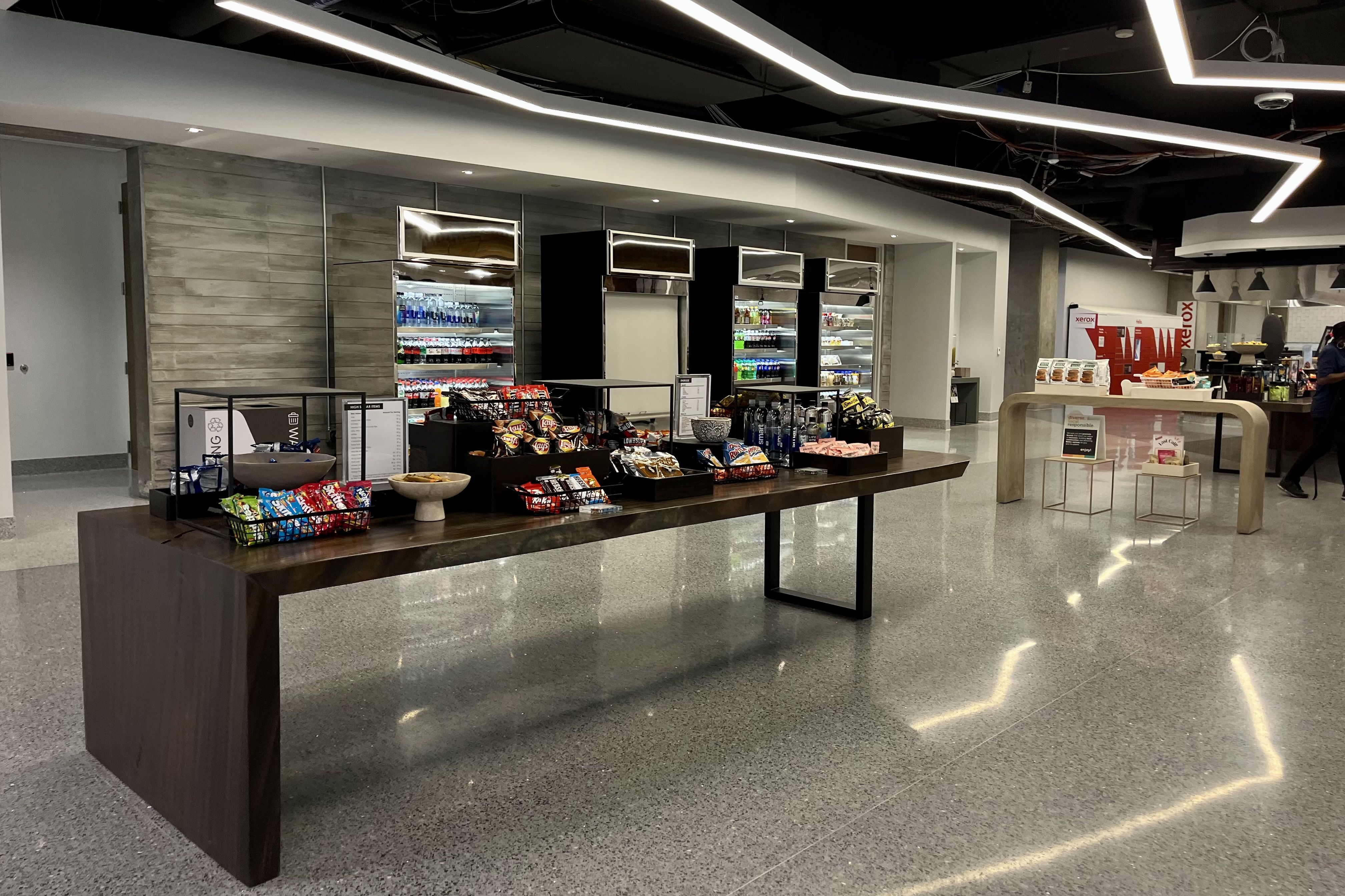 Modern snack and beverage area with a wooden table displaying chips, candy, and bottled water, three glass refrigerators stocked with drinks, and sleek overhead linear lighting in a polished space.