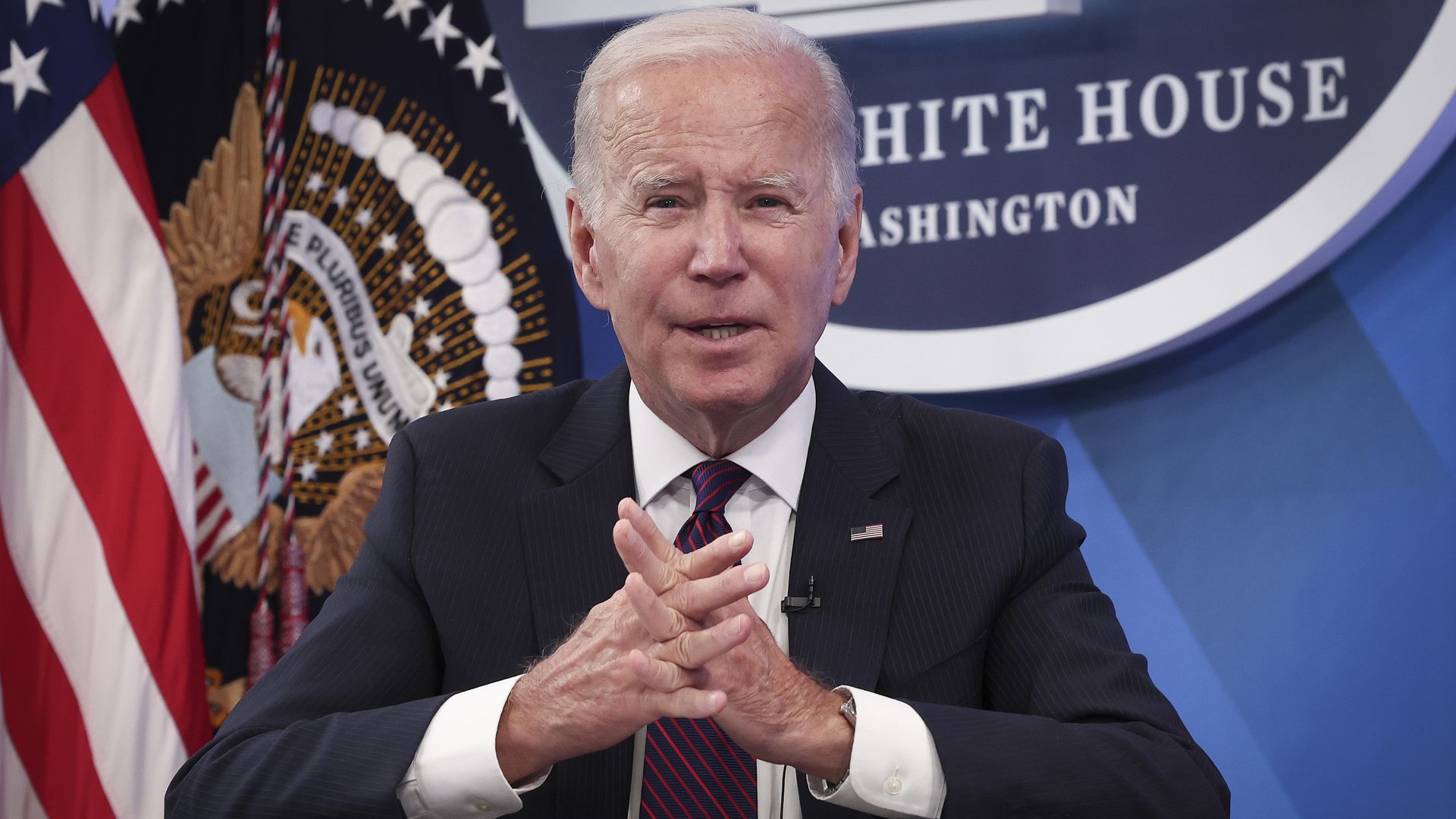 President Joe Biden sits at a desk delivering remarks in from of a White House sign and an American flag.