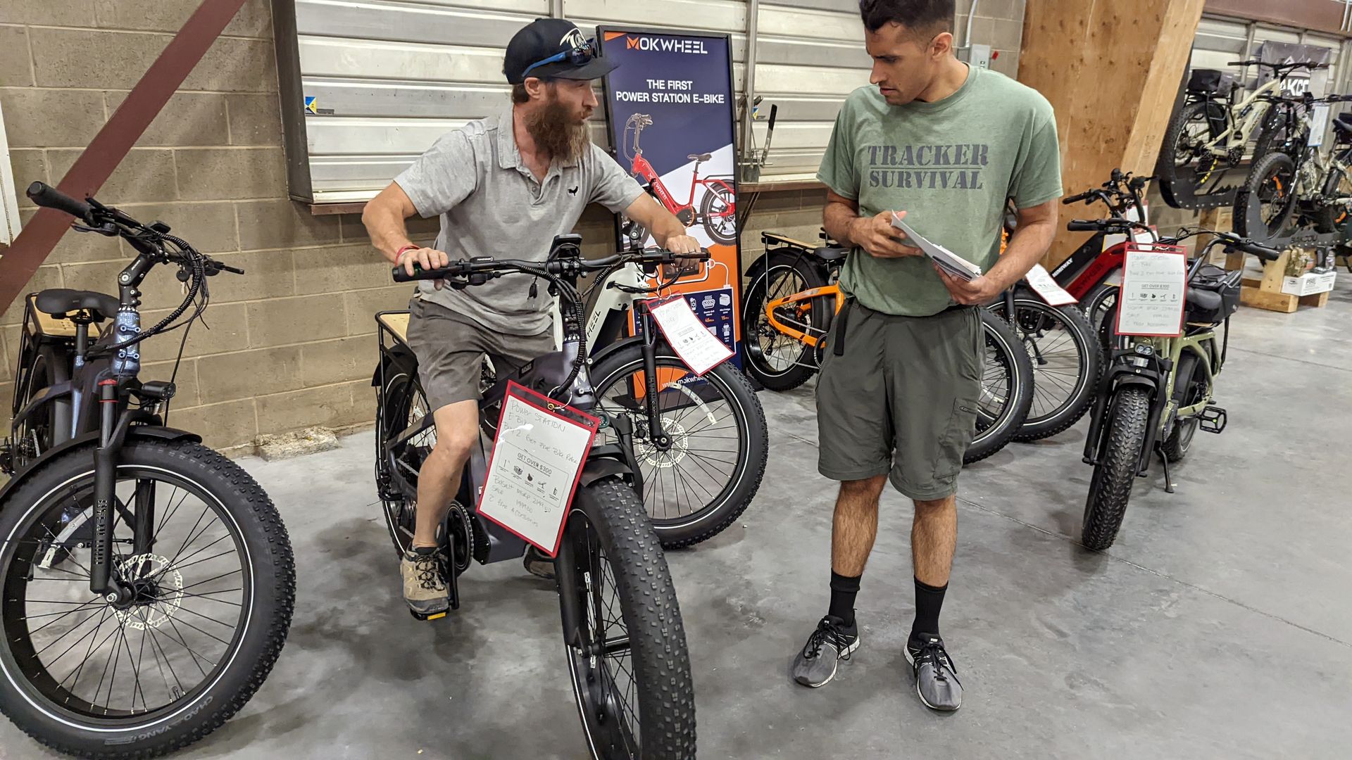 A man sits on an ebike on in an exhibition hall while another man looks at it. 