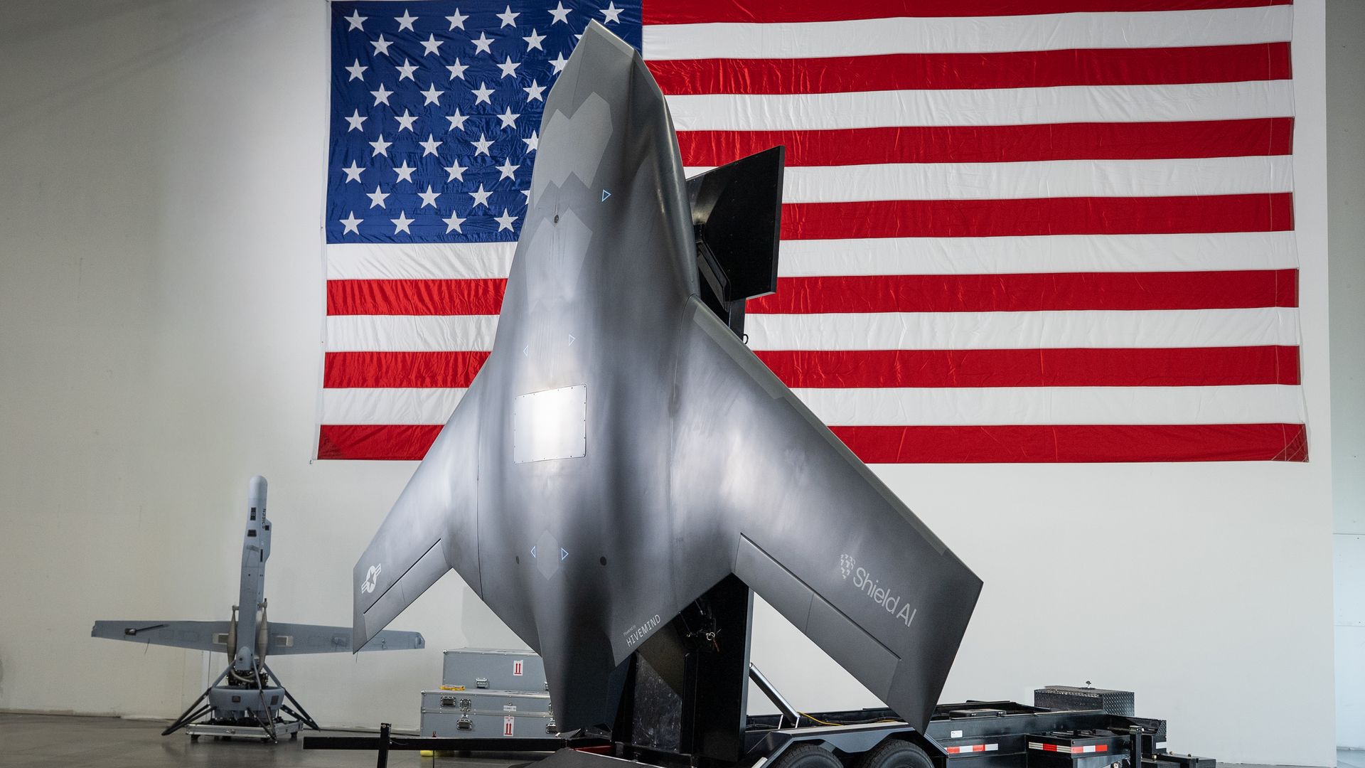 Black stealth drone named Hyemind Shield AI on wheels inside a hangar, with a large U.S. flag hanging on the wall behind it.