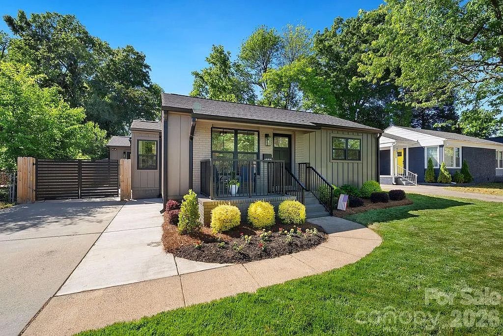 Single-story modern suburban home with gray siding and black railings on the front porch. A curved walkway leads to a landscaped bed with yellow shrubs; left driveway with a gate, green lawn and blue sky.e