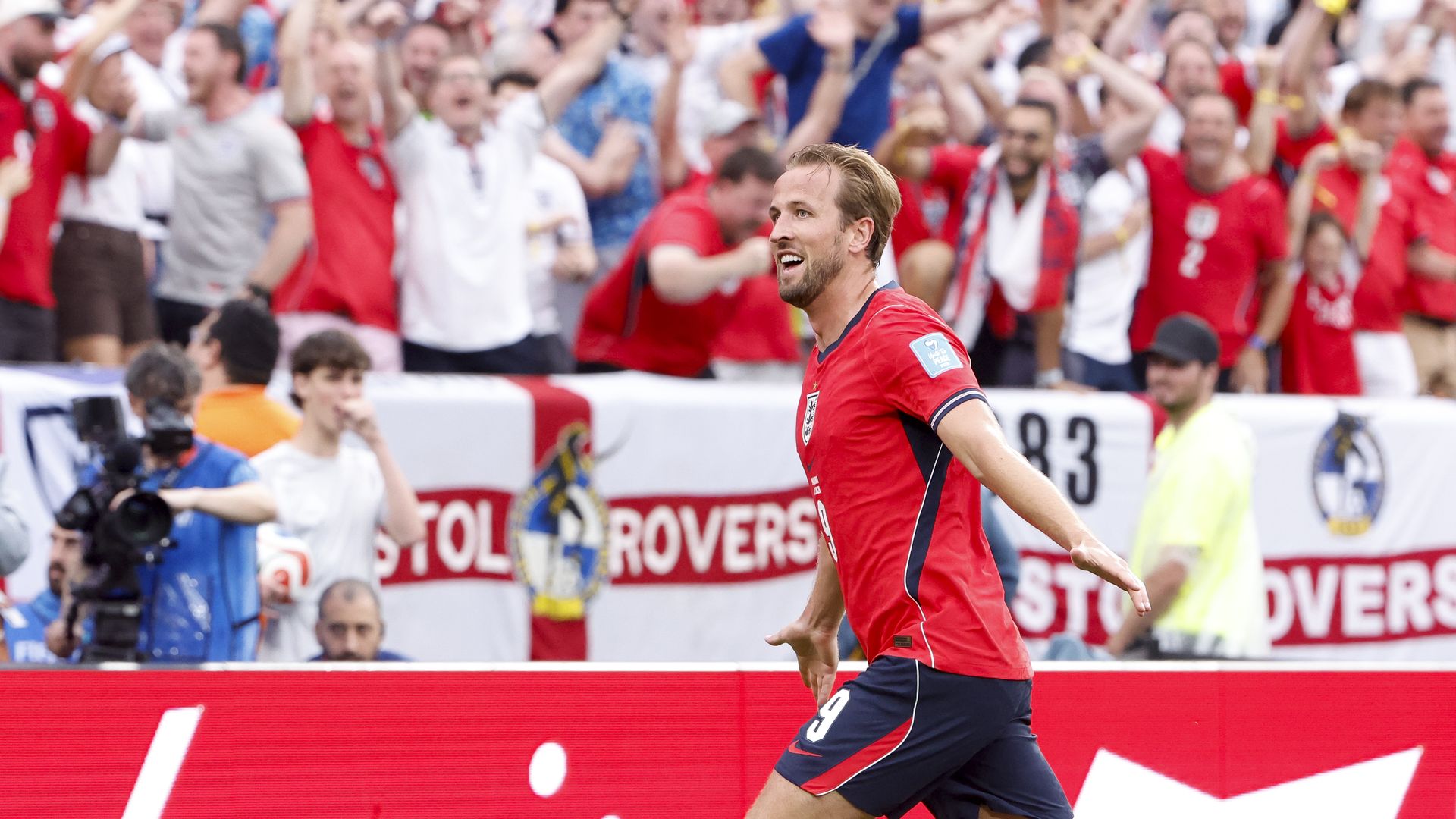 A man wearing a red soccer jersey smiles with his arms partially raised in the air. In the background, a crowd of spectators dressed in red and white cheers from the stadium stands. 