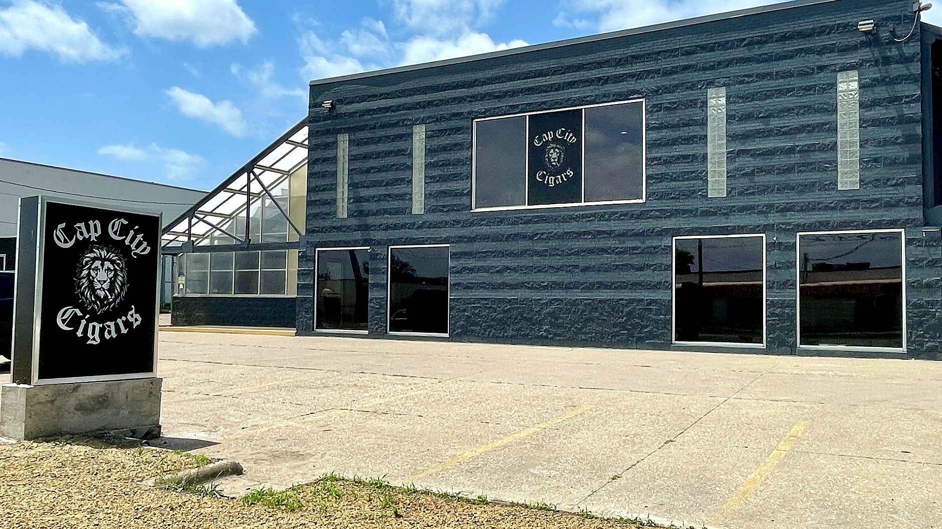 Dark blue, textured two-story building with large windows. A tall black sign reads "Cape City Cigars" with a lion emblem; a glass walkway attaches the structure. Clear blue sky with a few clouds.