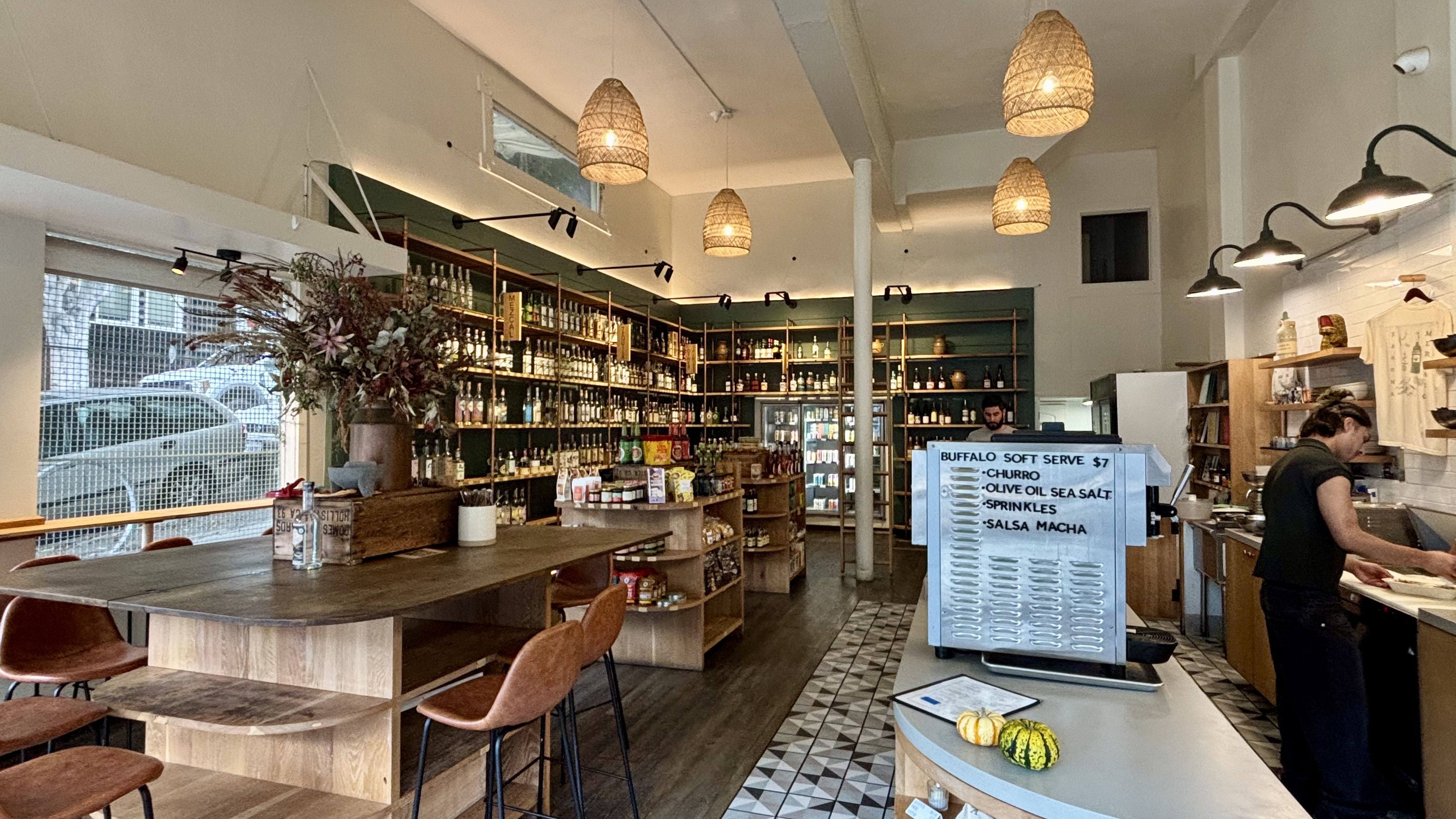 Cozy cafe interior with wooden tables, hanging wicker lamps, shelves stocked with bottles and goods, a staff member working behind a counter with a soft serve machine listing flavors.