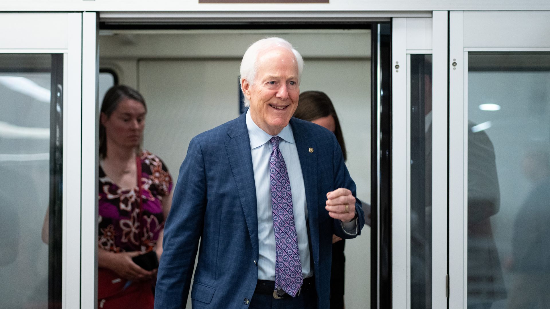 Smiling Texas Sen. John Cornyn in a blue suit and purple patterned tie walking out of subway doors with a "DO NOT HOLD SUBWAY DOORS" sign above him. Two people blurred in background.
