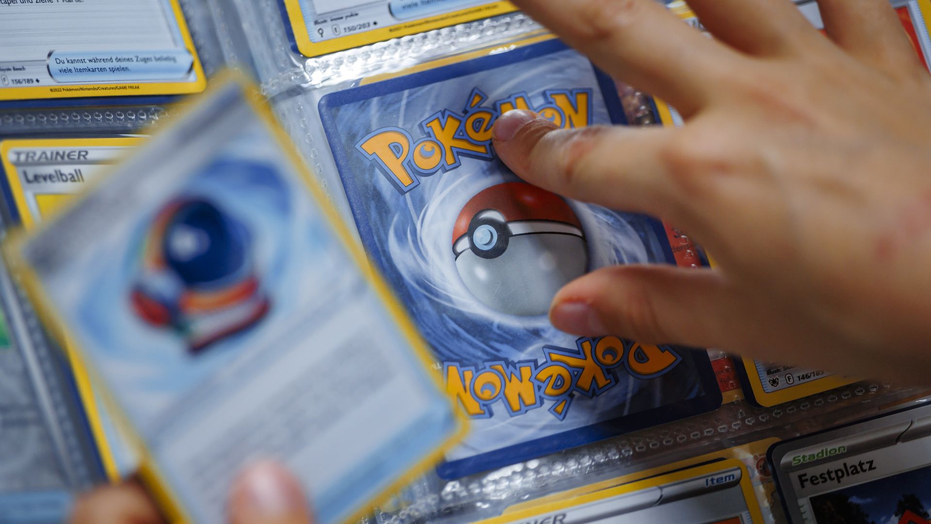 Person's hand holding and placing Pokémon trading cards in a binder with visible blue, yellow, and white card designs and a Poké Ball image.