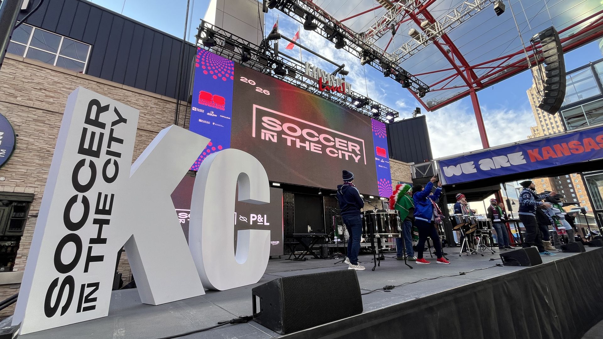 Outdoor stage setup at KC Live! downtown Kansas City with large white letters "KC" and text "SOCCER IN THE CITY" in black, performers on stage wearing scarves, and a large LED screen displaying "SOCCER IN THE CITY" under a blue sky.