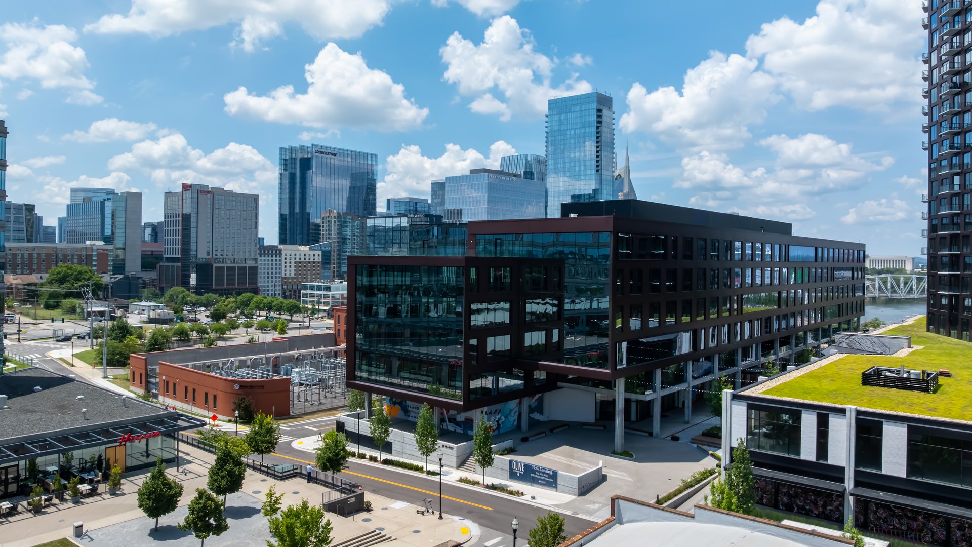 Blue-sky cityscape with white clouds and a cluster of glass office buildings; a tall blue-tinted tower stands center, plus a right-side high-rise with balconies, and city streets below.
